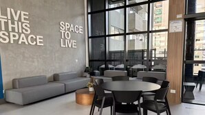 Et moderne loungeområde med en grå sofa, runde borde og stole, store vinduer samt et skilt med teksten 'LIVE THIS SPACE'.