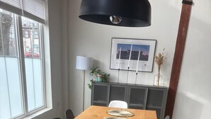 A long wooden table with woven placemats, black chairs, a black pendant light, a framed picture, and a cabinet with a plant.
