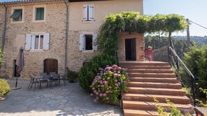 Un bâtiment en pierre avec un escalier menant à une porte, des places assises à l’extérieur et un jardin fleuri.
