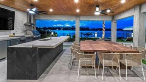 An outdoor kitchen with a dining area, a grill, and a view of a lake at dusk.