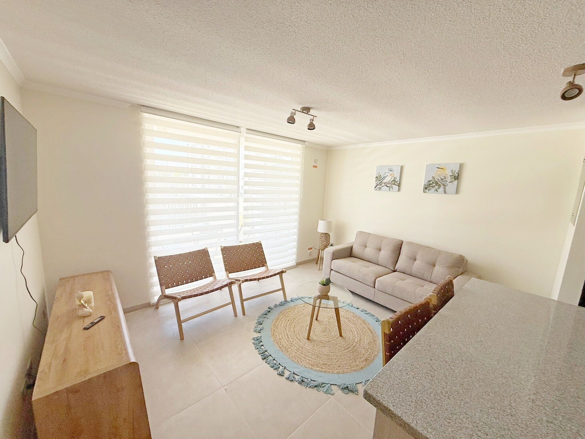 Ein Wohnzimmer mit einer Couch, zwei Sesseln, einem Couchtisch, einem Fernseher und einem Fenster mit Jalousien.