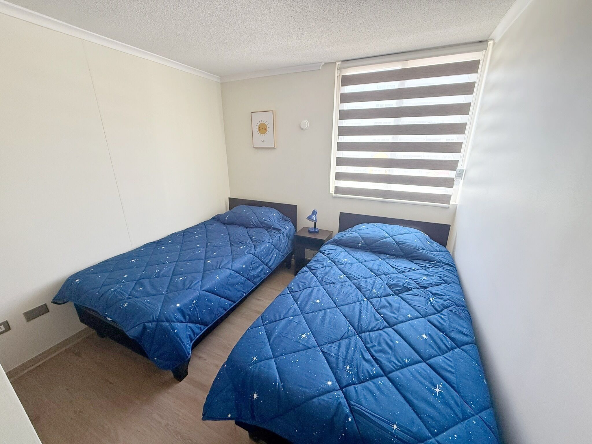 Une chambre avec deux lits simples, une table de chevet avec une lampe, une fenêtre avec des stores et une horloge murale.