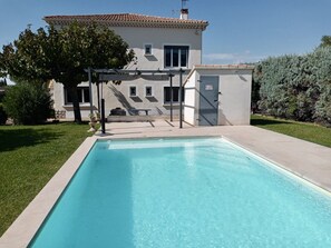 A rectangular swimming pool with a tiled deck area, a pergola, and a house with a tiled roof in the background. - La Maison de Gari, spacious house with swimming pool and beautiful exterior (Taillades)