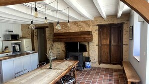 Une cuisine rustique avec une table en bois, des chaises, des ampoules suspendues, un poêle en briques et un banc.