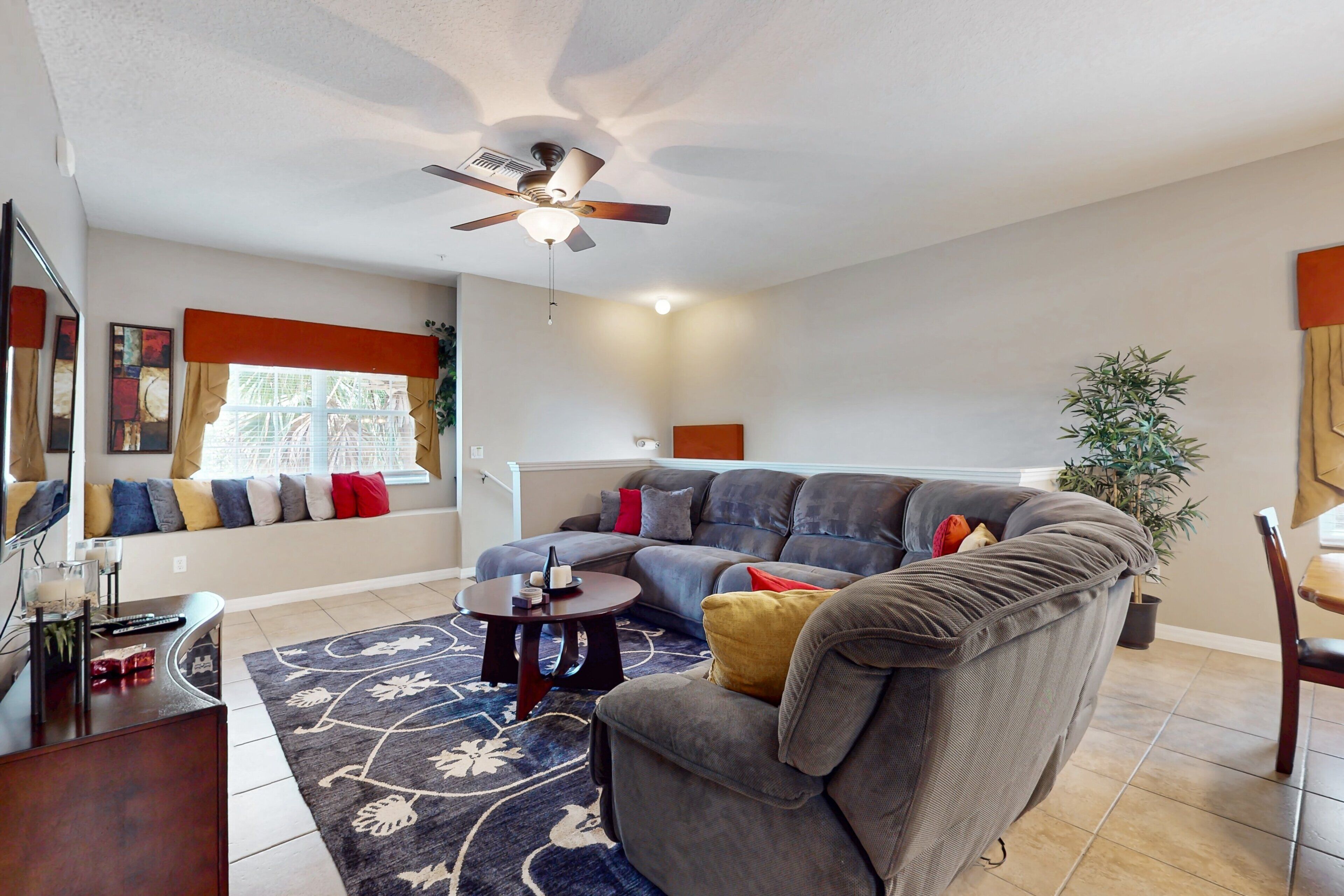 A living room with a sectional sofa, a ceiling fan, a dining area with a table and chairs, and a potted plant.
