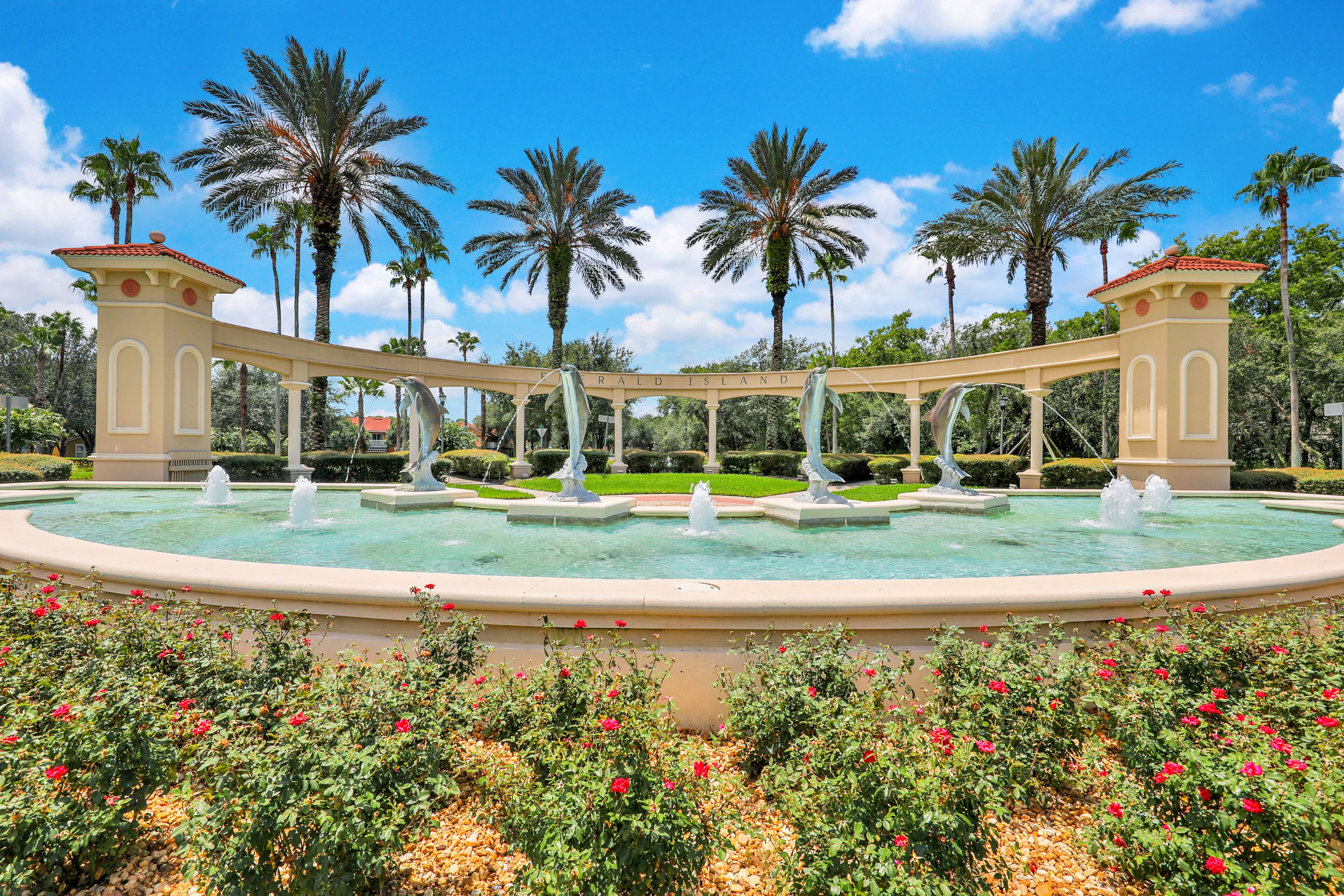 A circular fountain with statues, surrounded by red flowers and palm trees.