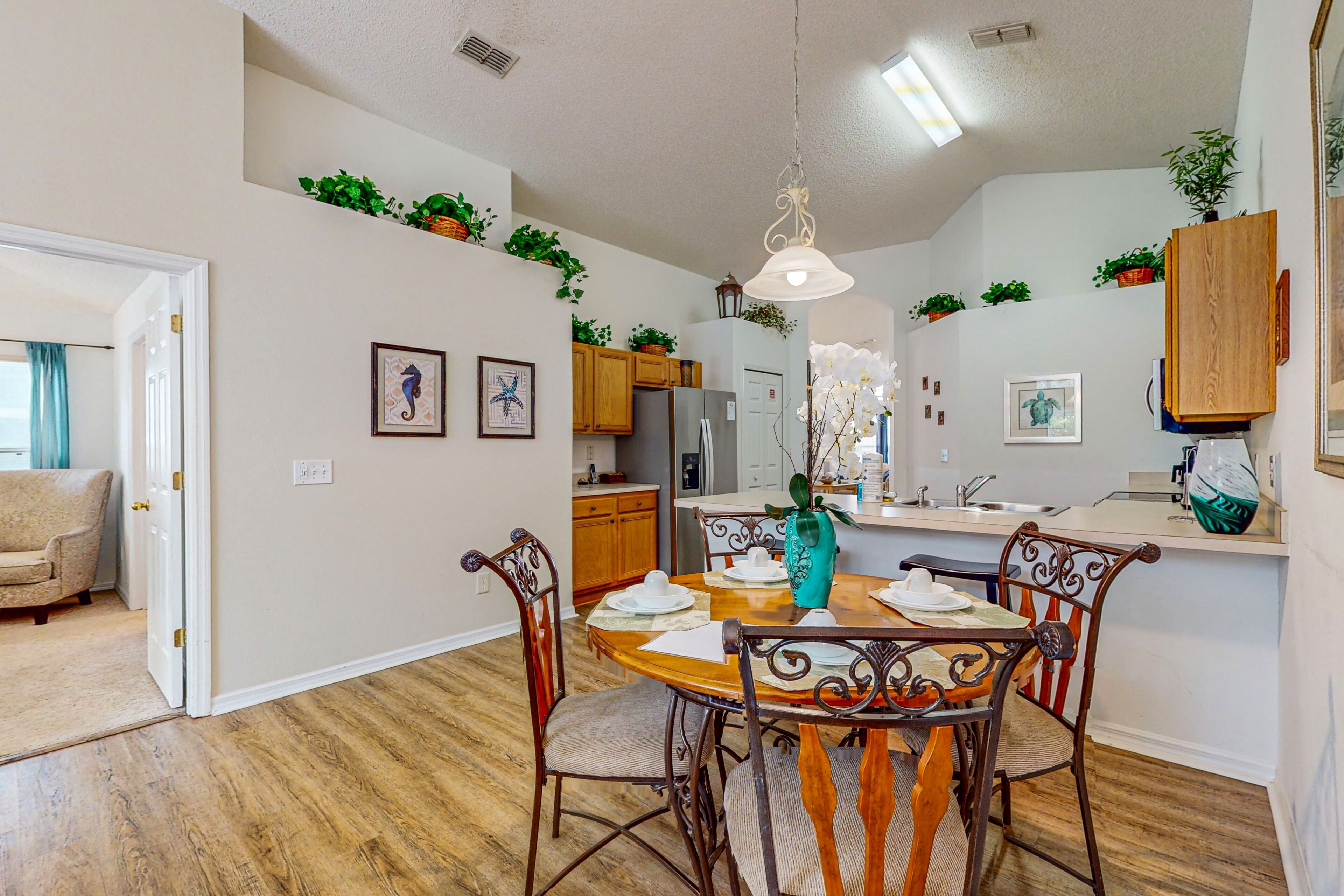 A dining area with a wooden table, chairs, and a vase. There are plants on the walls and a kitchen in the background.