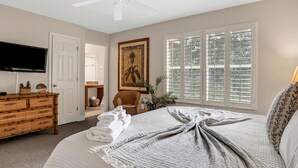 Ein Schlafzimmer mit einem Bett, einem Fernseher, einem Deckenventilator, einem Fenster mit Jalousien, einem Gemälde und einem Korbmöbel-Kommoden.