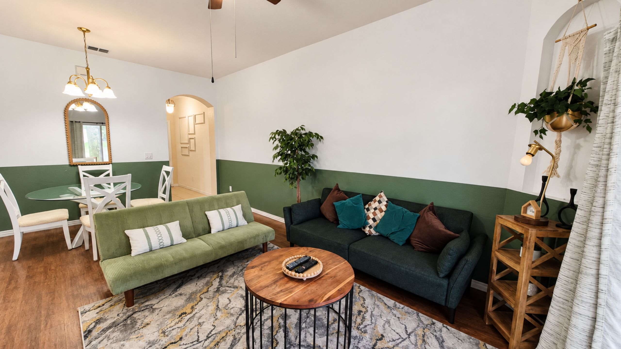 Uma sala de estar com um sofá verde, uma mesa de centro redonda e um espaço de jantar com mesa de vidro e cadeiras brancas.