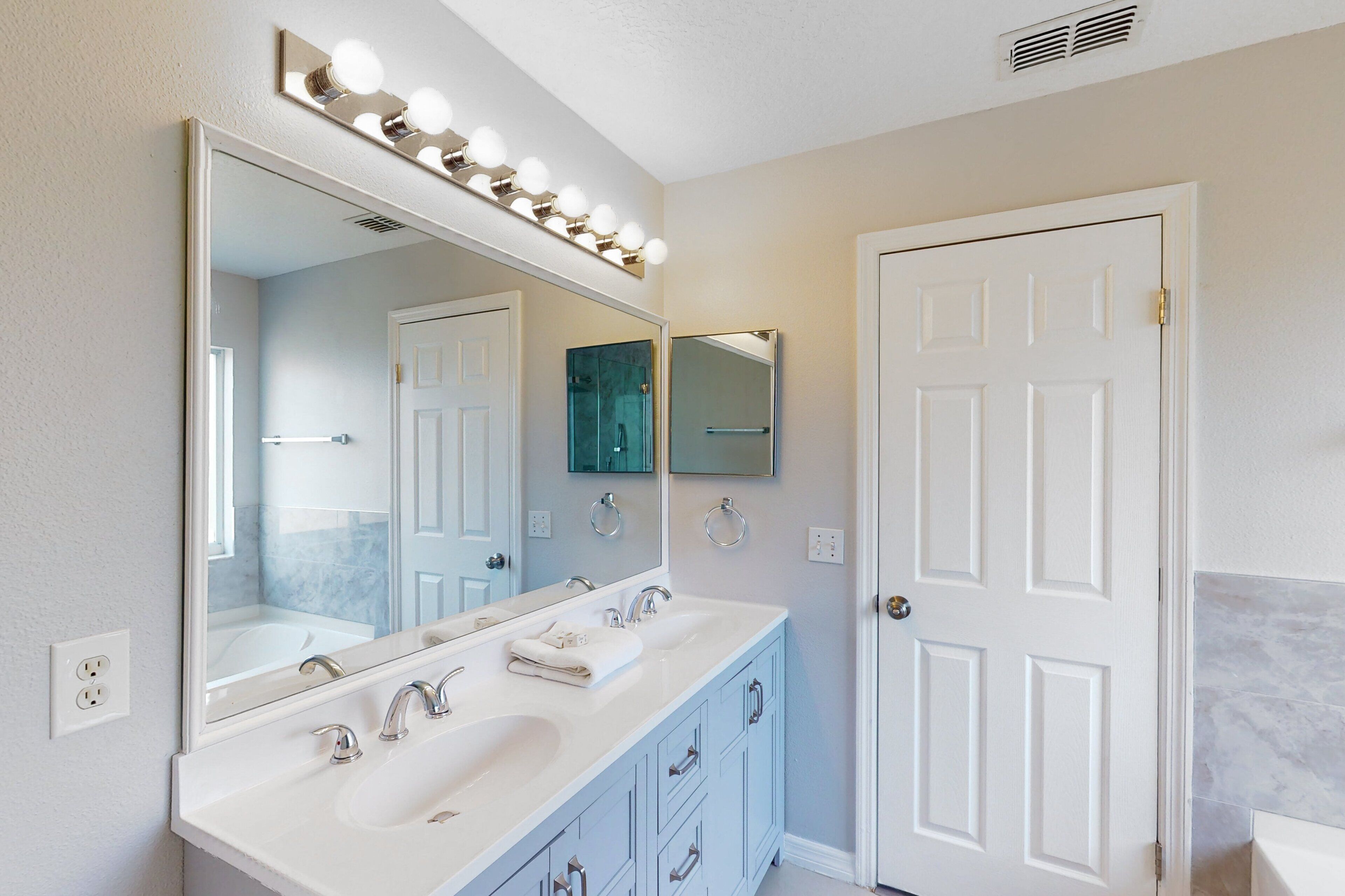 A bathroom with a large mirror, two sinks, vanity cabinets, and a door.