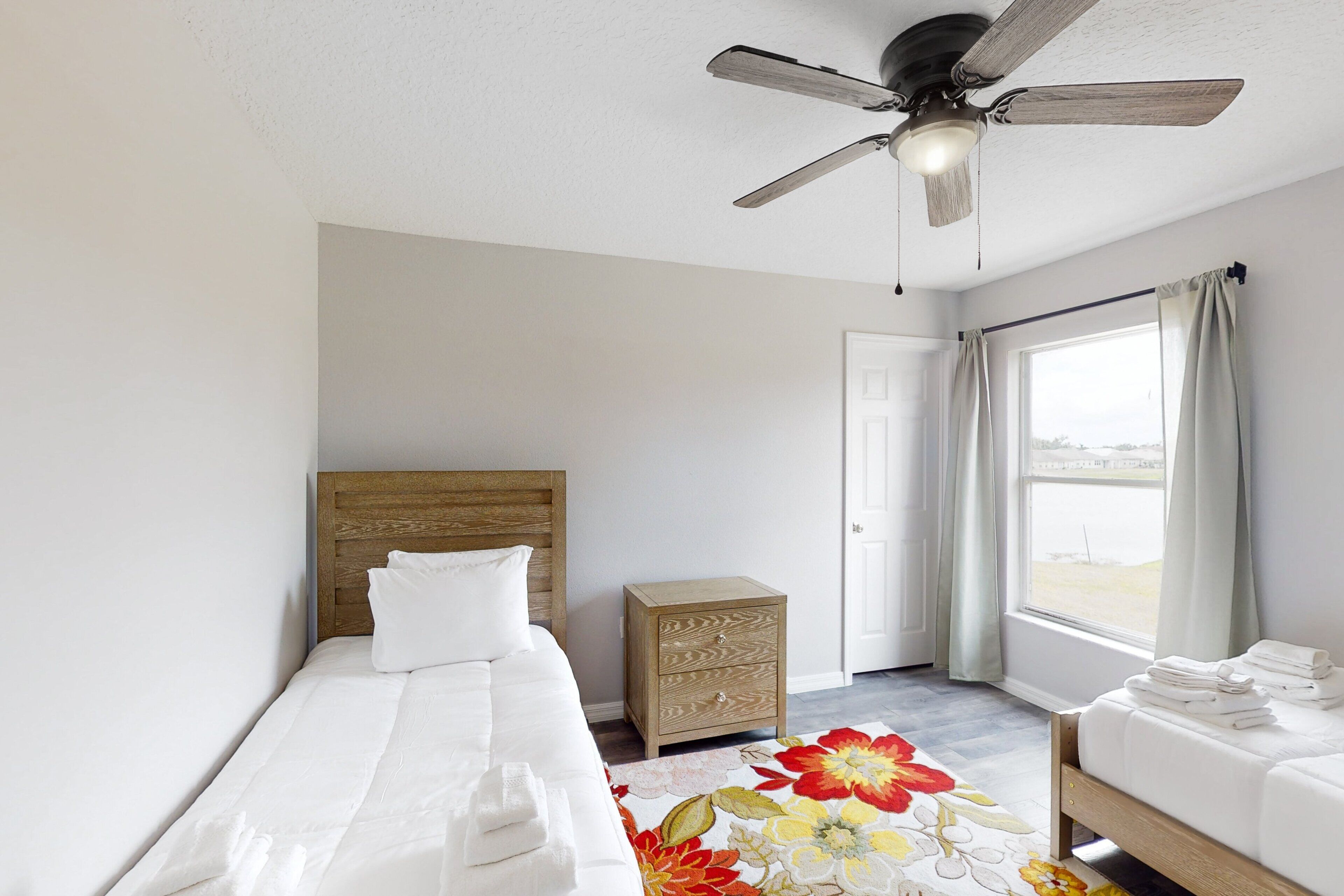A bedroom with a bed, a nightstand, a ceiling fan, a window with curtains, and a view.