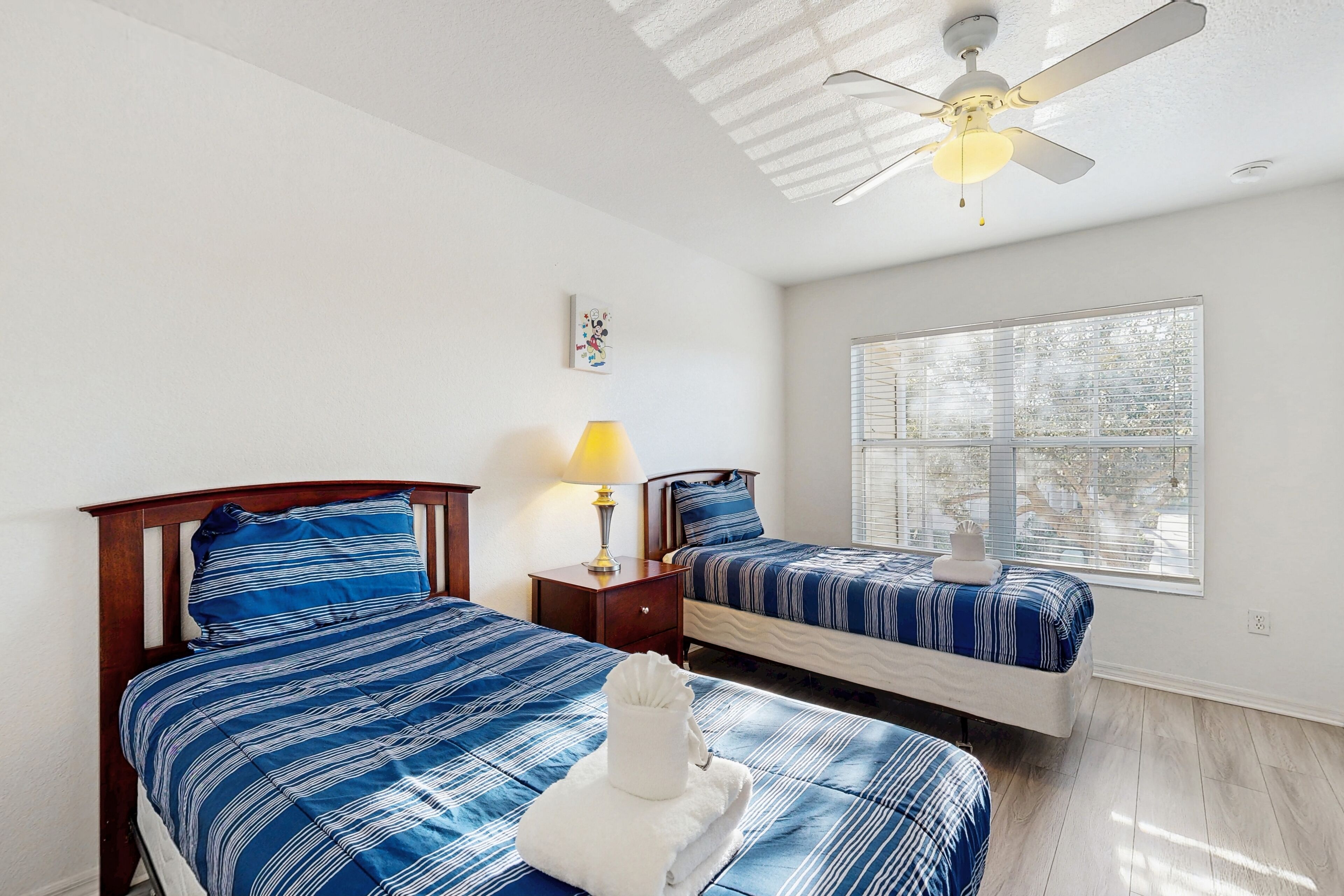 A bedroom with two beds, a ceiling fan, a window with blinds, and a bedside table with a lamp.