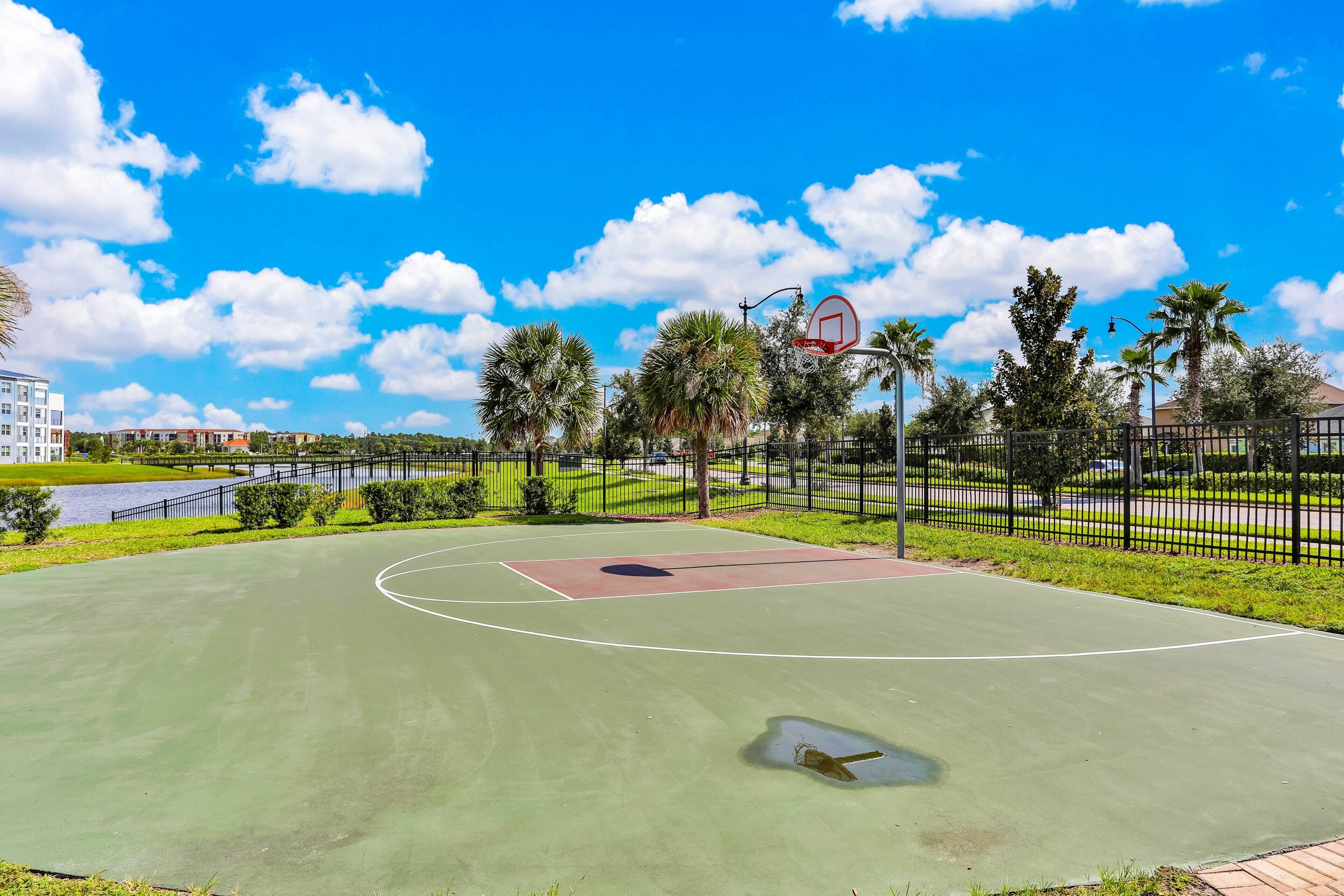 An outdoor basketball court with a clear sky, palm trees, and a fence.