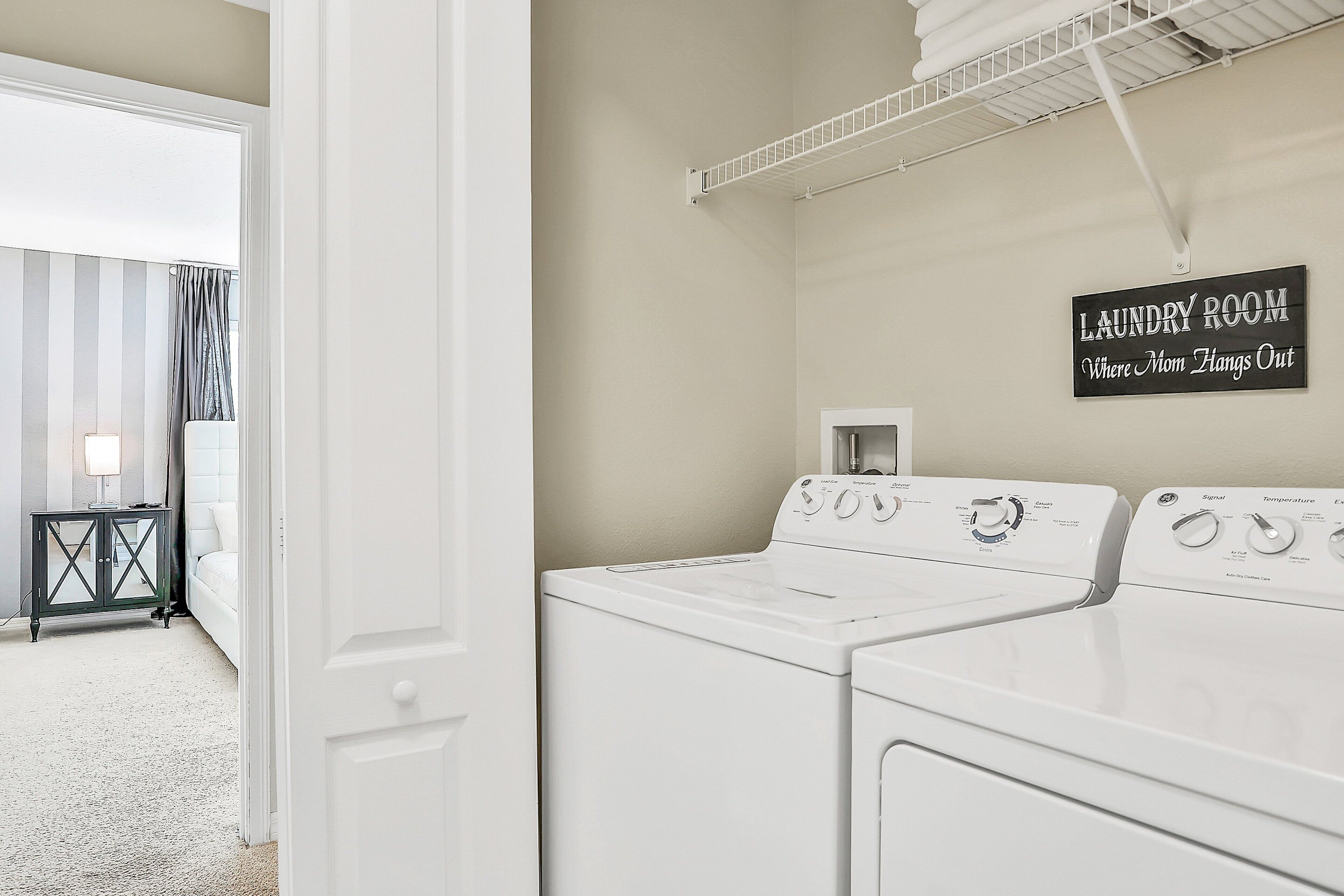 A laundry room with a sign reading 'Laundry Room Where Mom Hangs Out,' a white washing machine, and a drying rack.