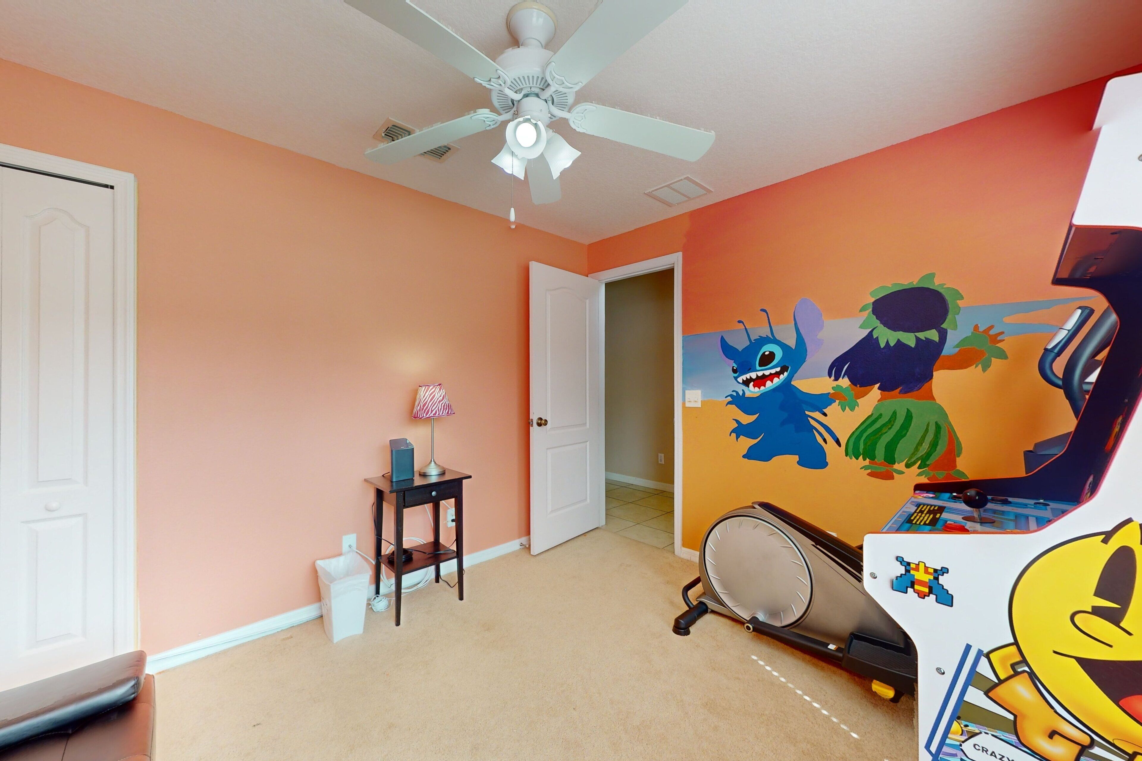 A room with a ceiling fan, a door, a small table with a lamp, and a wall with colorful cartoon characters.