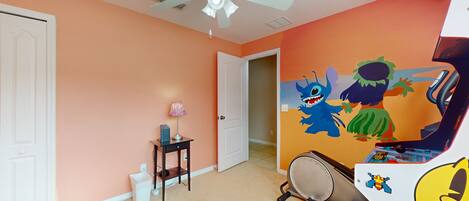A room with a ceiling fan, a door, a small table with a lamp, and a wall with colorful cartoon characters.