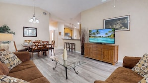 Una sala de estar con televisor, sofá, mesa de comedor, sillas y una cocina al fondo.