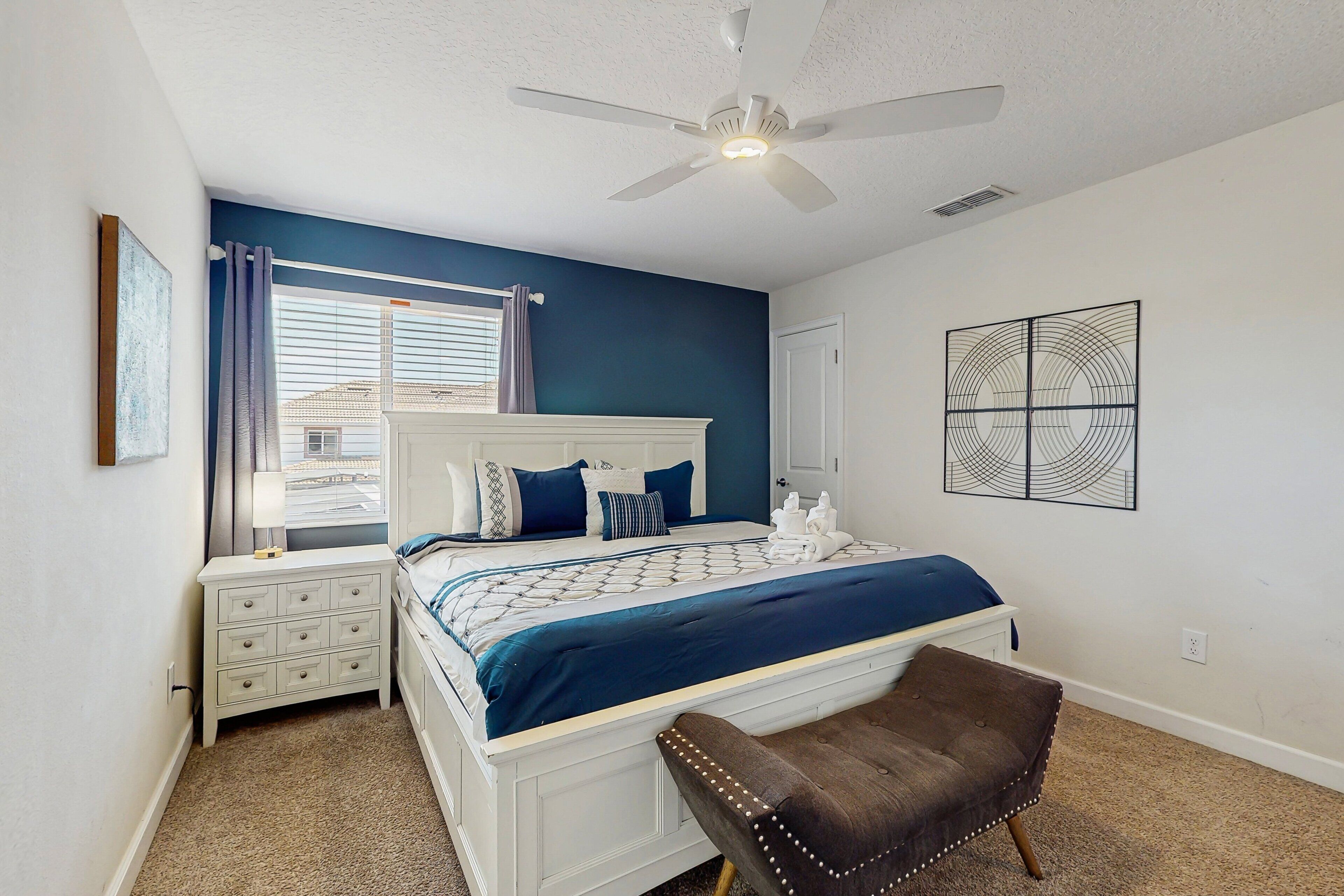 A bedroom with a bed, nightstand, bench, ceiling fan, window with curtains, and a wall art piece.