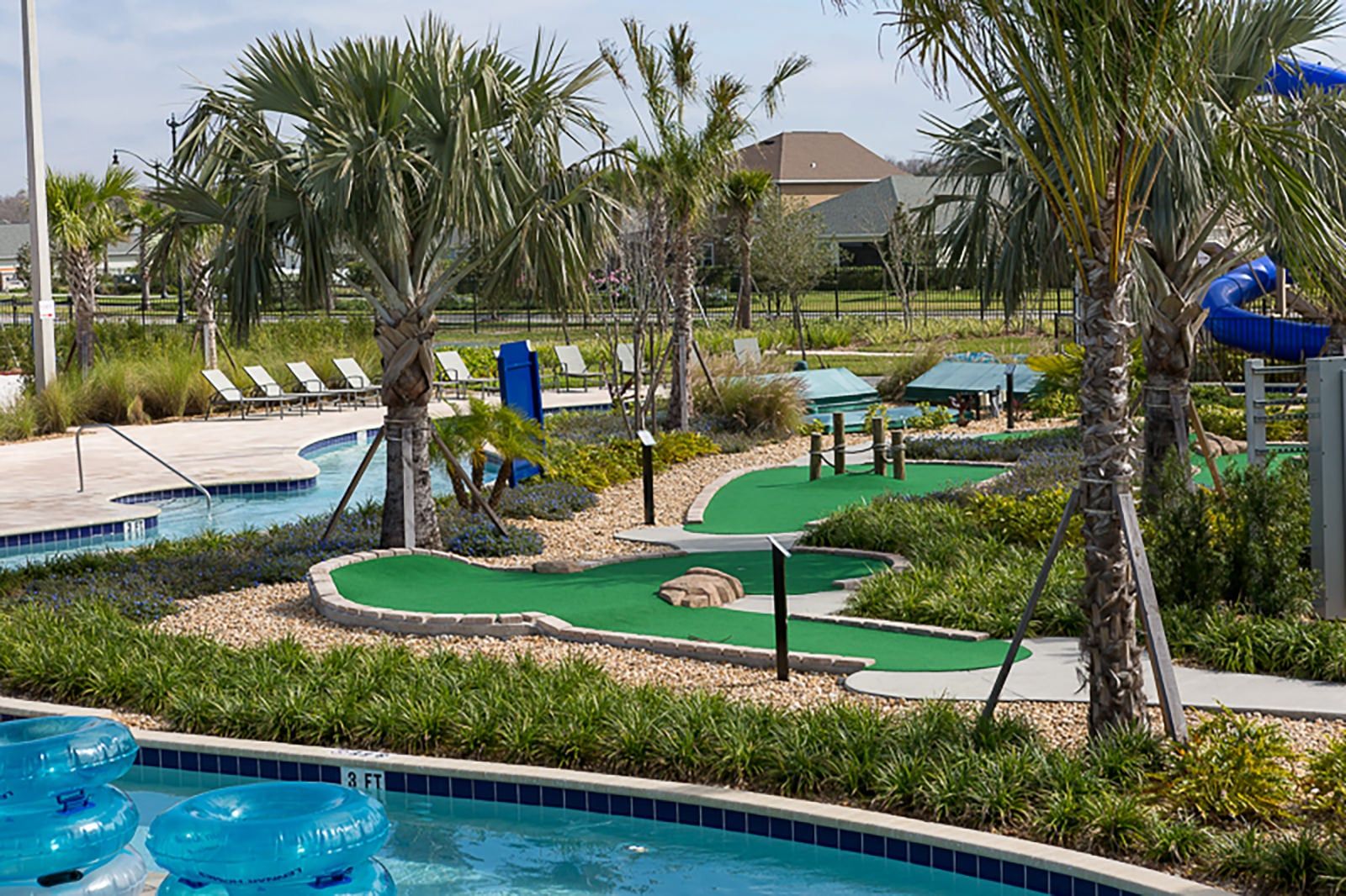 A pool area with a putting green, lounge chairs, palm trees, and a building in the background.