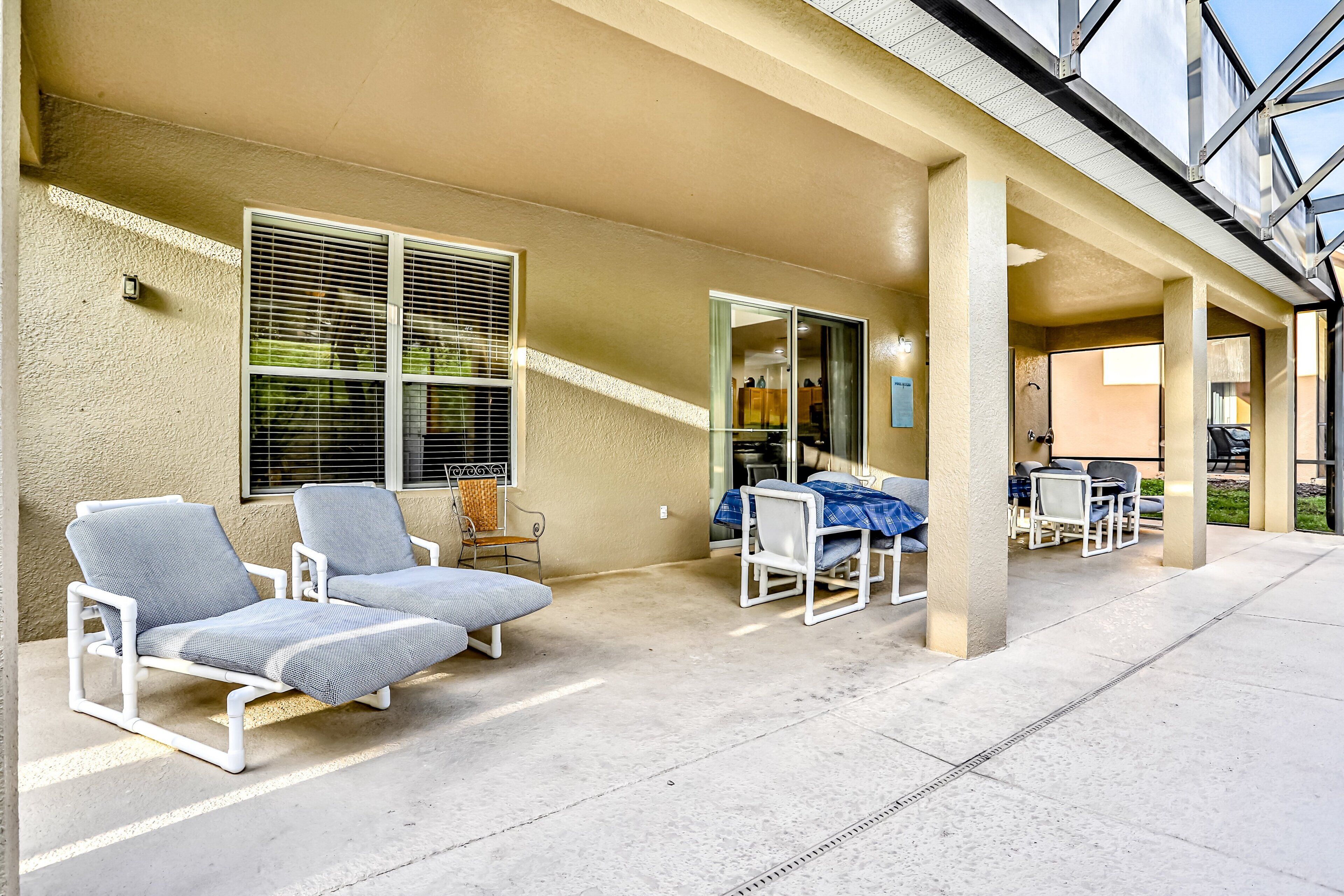 A covered outdoor seating area with lounge chairs and a table, a window with blinds, and a view of the interior.