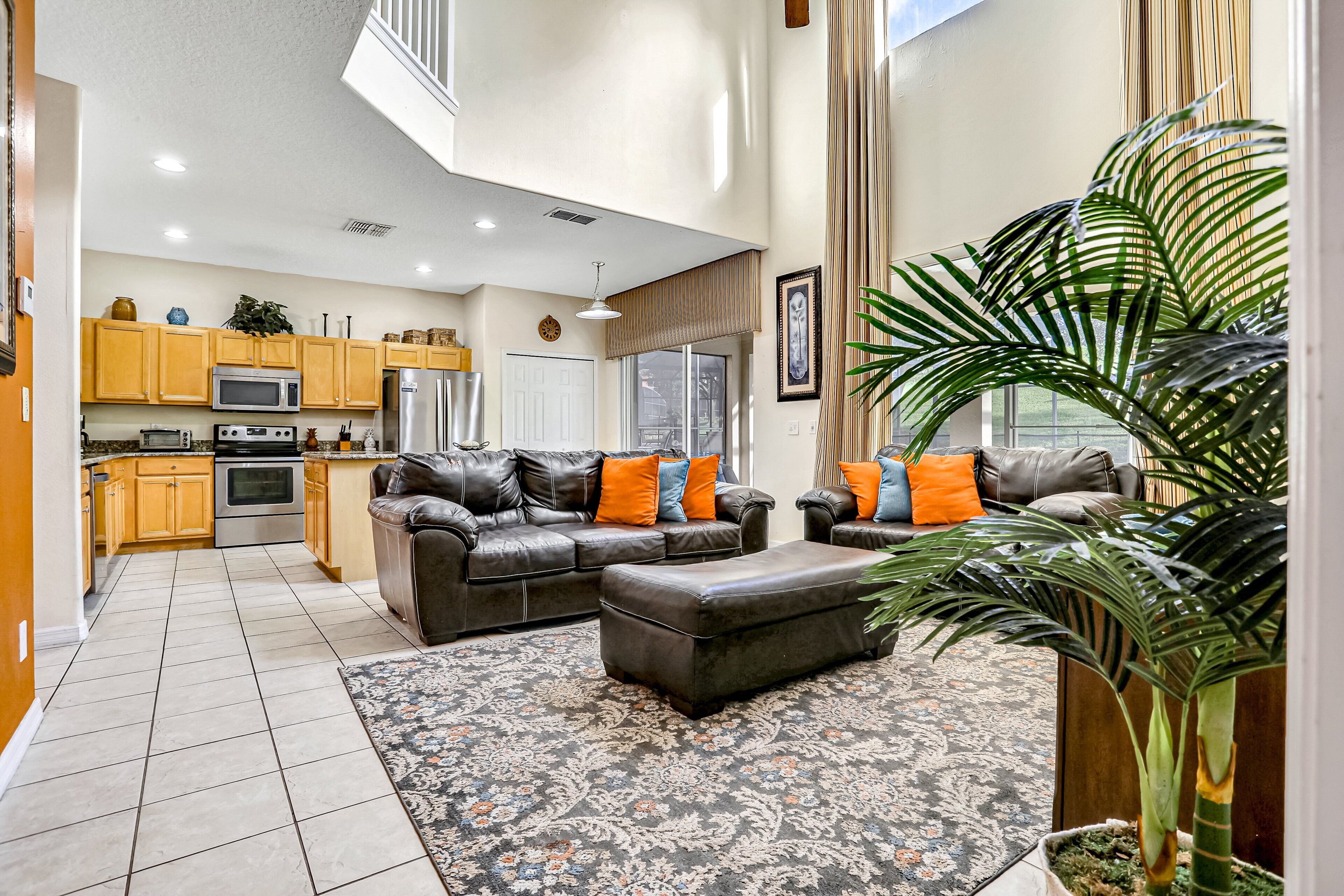 A spacious living area with a sofa and ottoman, a kitchen with wooden cabinets, a large window, and a potted plant.
