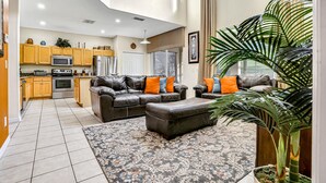 Een ruime woonkamer met een bank en voetenbankje, een keuken met houten kastjes, een groot raam en een potplant.