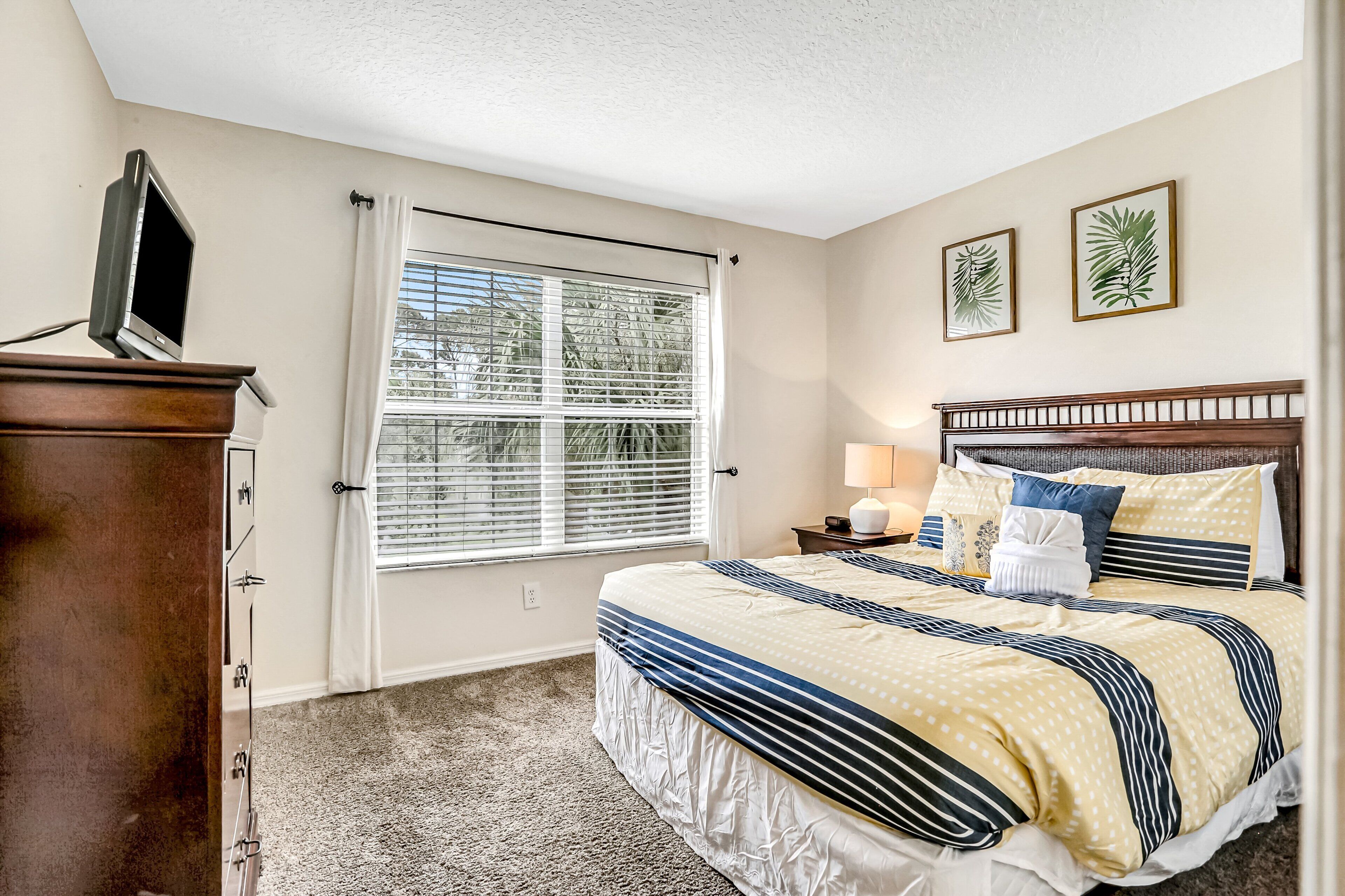 A bedroom with a bed, a nightstand, a lamp, a dresser, a window with blinds, and two wall art pieces.