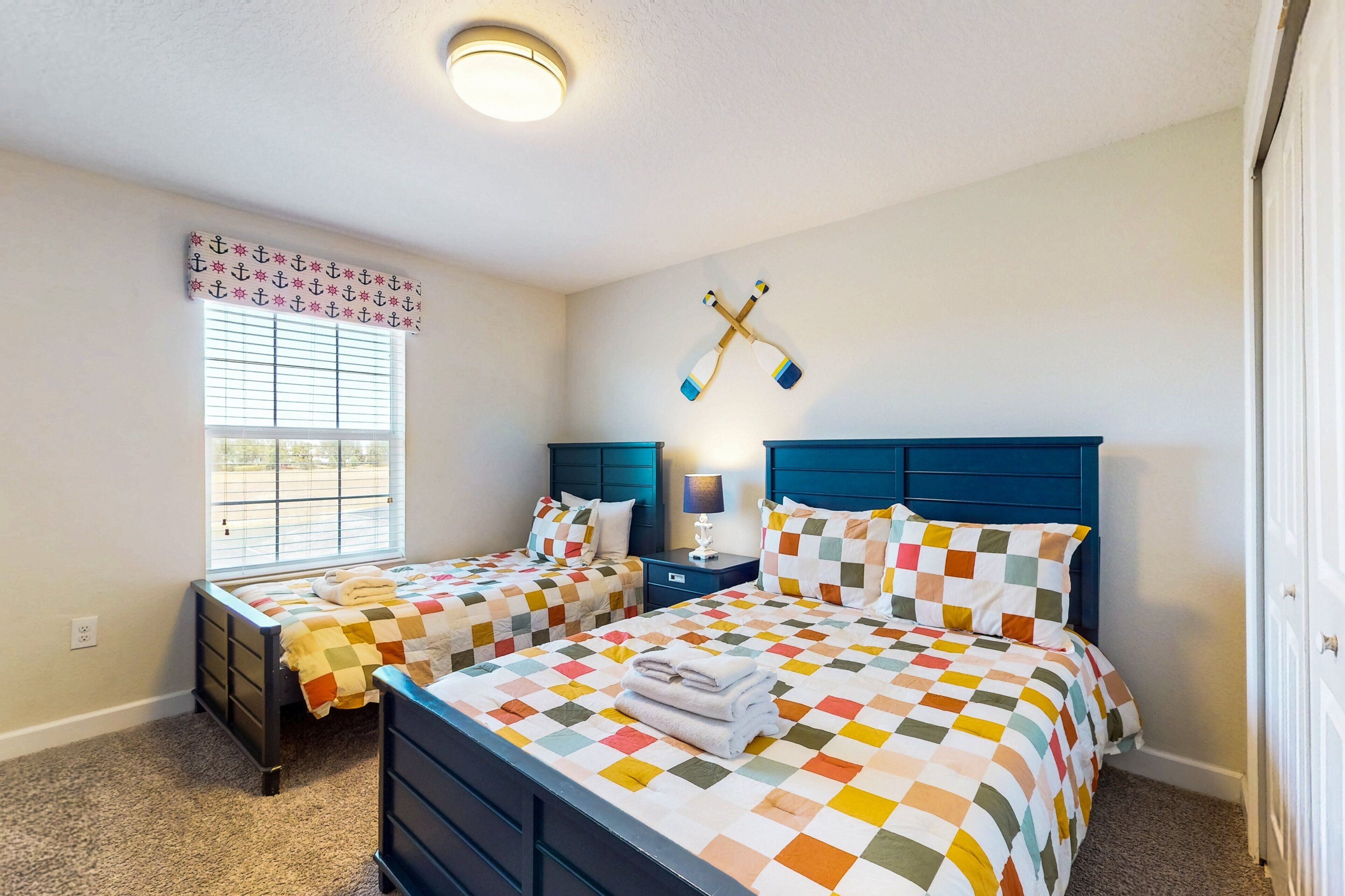 A bedroom with two beds, a window with blinds, a wall-mounted cross, and a nightstand with a lamp.