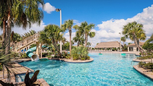 Un área de piscina en un resort, con un tobogán de agua, palmeras y un edificio con techo de paja.