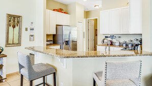 A kitchen with white cabinets, a granite countertop, and a stainless steel refrigerator. There are two bar stools and a painting on the wall. - Dog-friendly Home w/ Private Pool, High-speed Wifi, and Central AC (Kissimmee)