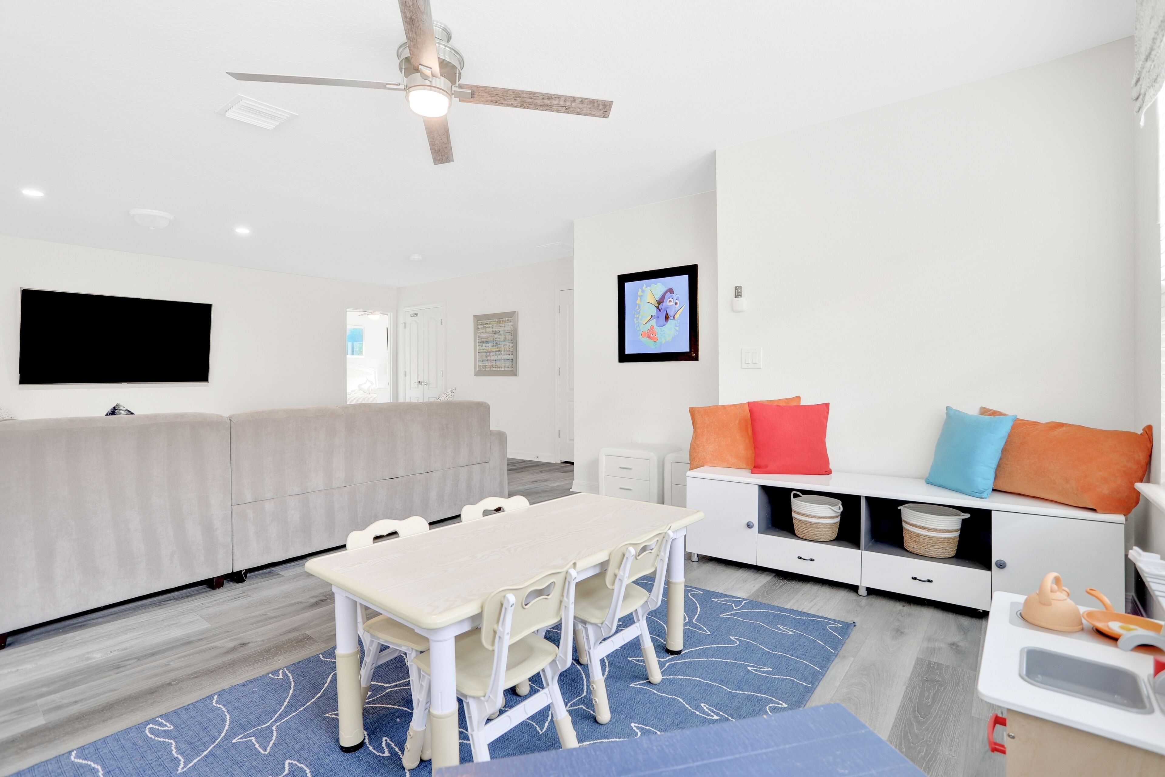 Uma sala de estar moderna com um sofá branco, uma mesa de jantar com cadeiras, um ventilador de teto, uma televisão de tela plana e uma kitchenette com pia e fogão.