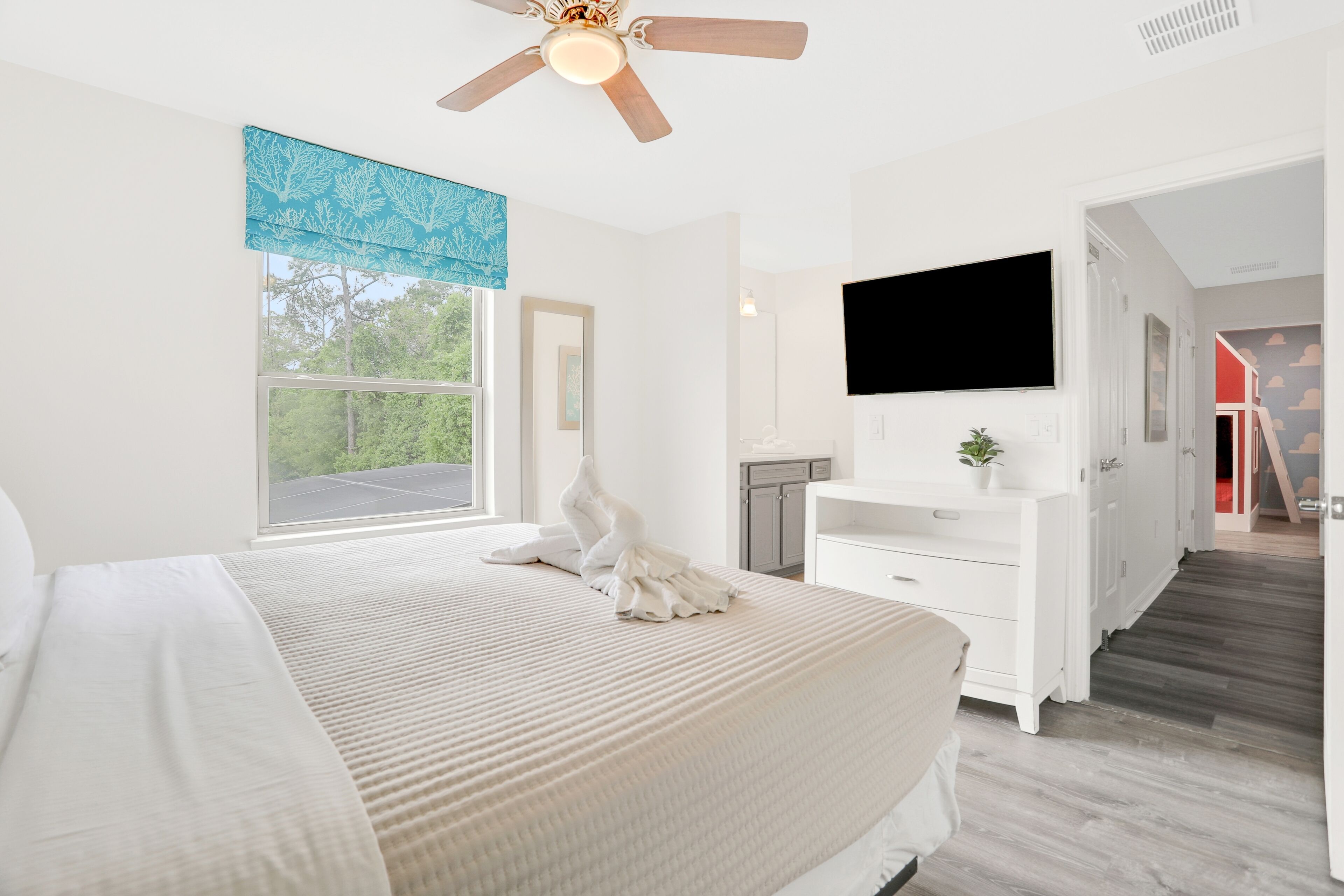 A bedroom with a large bed, a ceiling fan, a TV mounted on the wall, and a window with a view of trees.