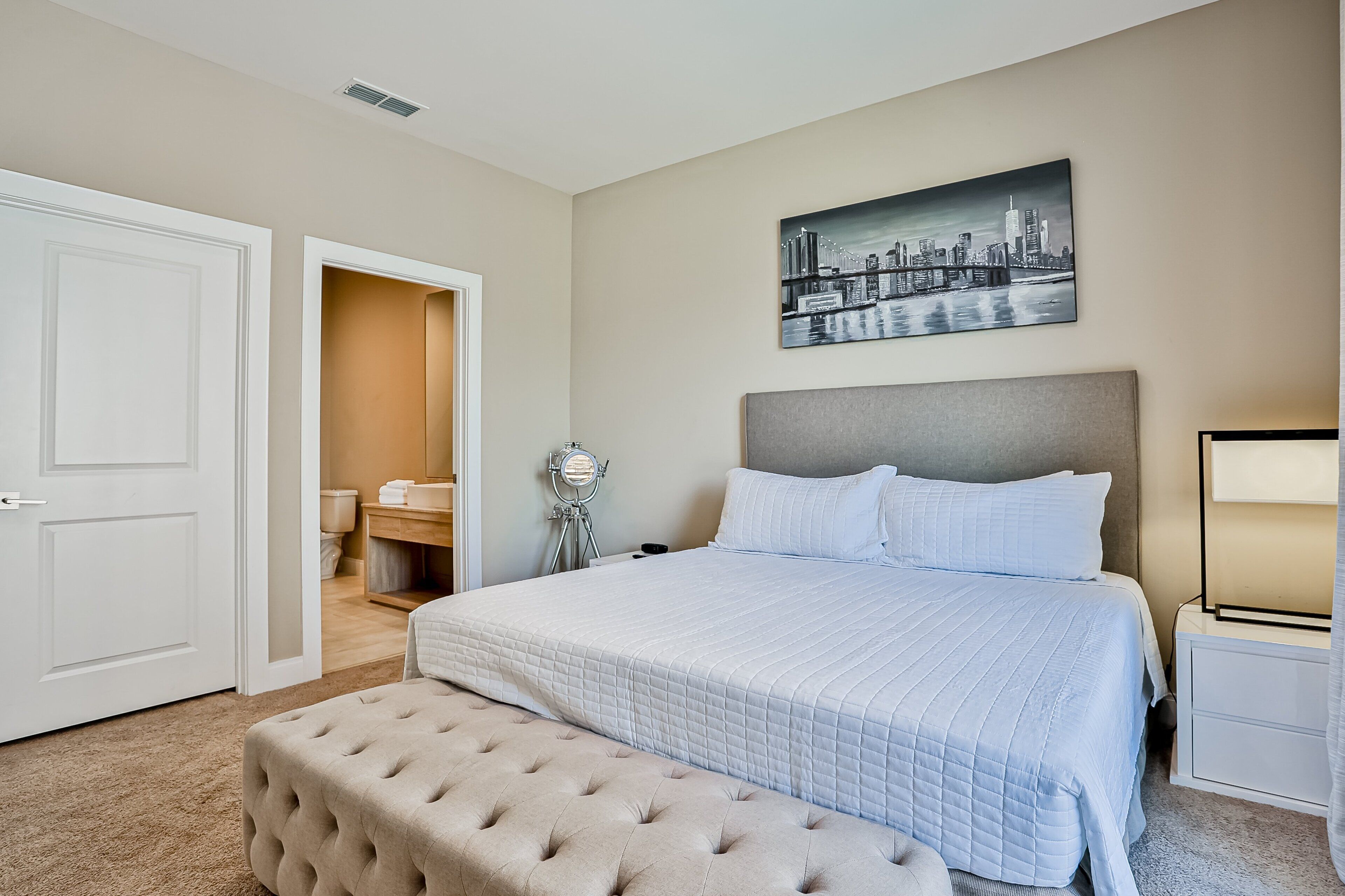 A bedroom with a bed, a bench, a nightstand, a mirror, and a cityscape painting on the wall.