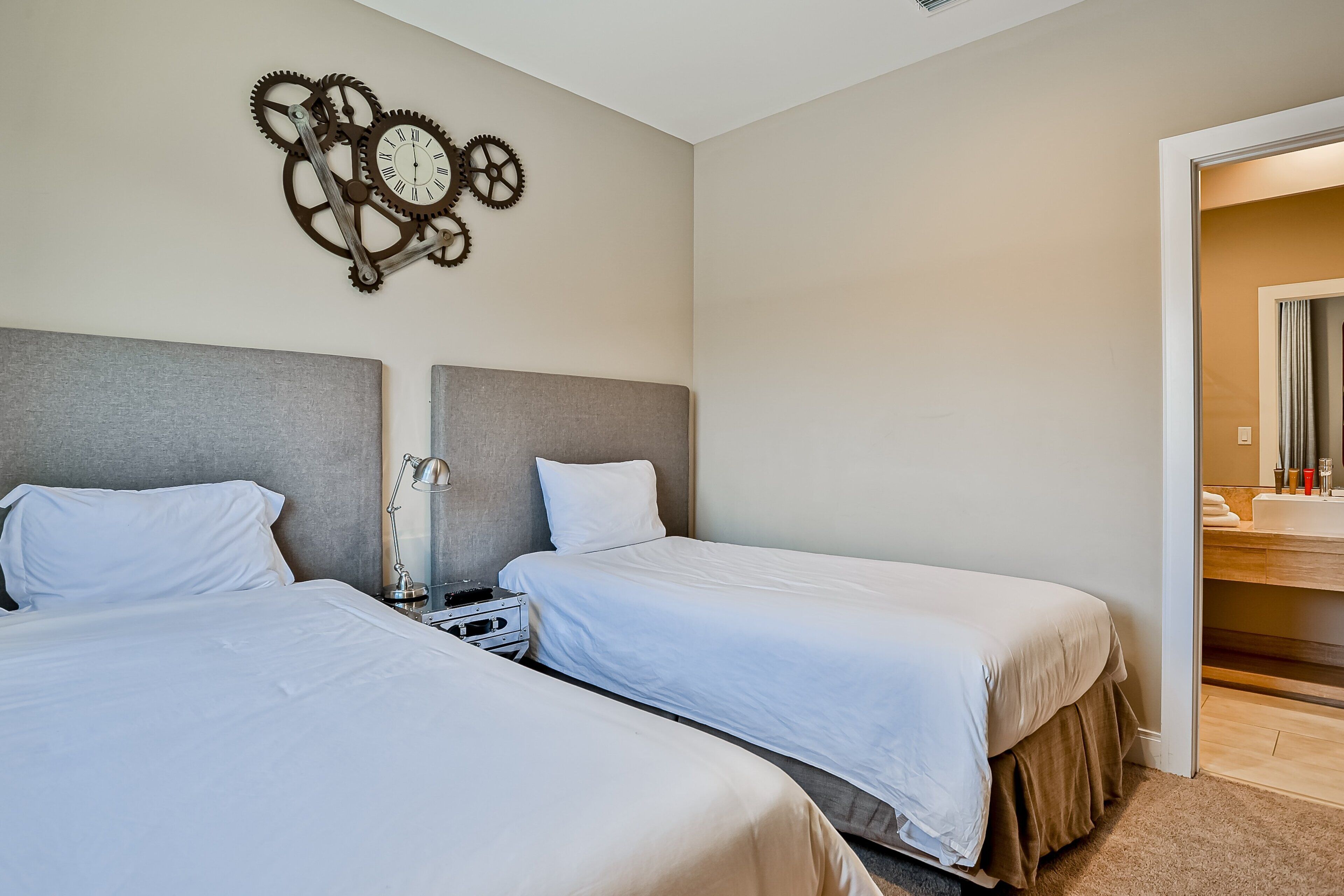 Quarto de hotel com duas camas, um relógio com engrenagens na parede e um banheiro visível por uma porta aberta.