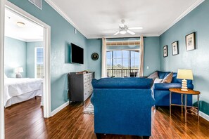 Quarto com cama, sofá azul, televisão, ventilador de teto e janela com vista.