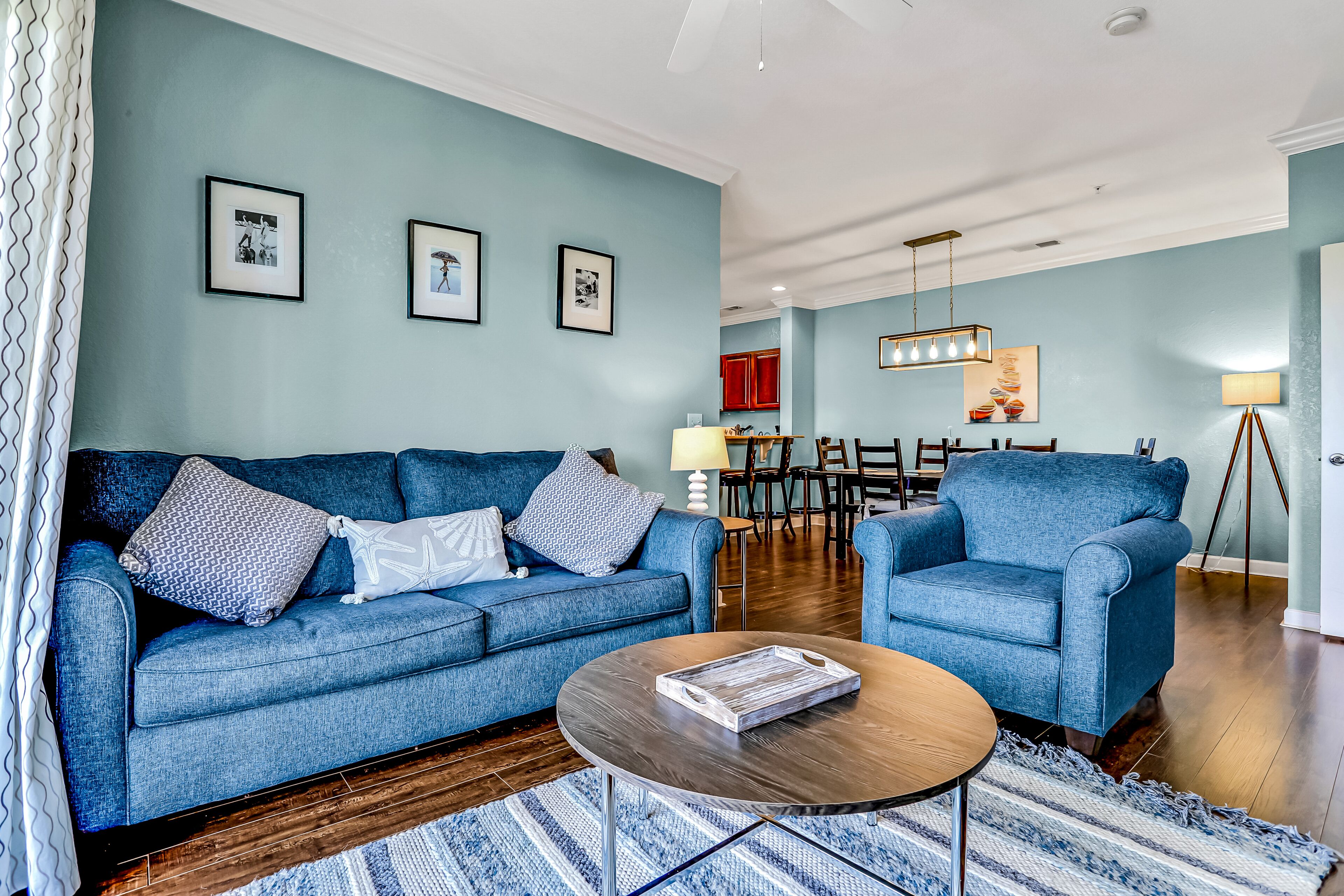 Un salon avec un canapé bleu, un fauteuil bleu, une table basse et, au fond, un coin repas.