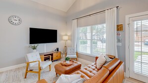 Een moderne woonkamer met een flatscreen televisie, een leren bank, een witte fauteuil en een groot raam met zonwering.