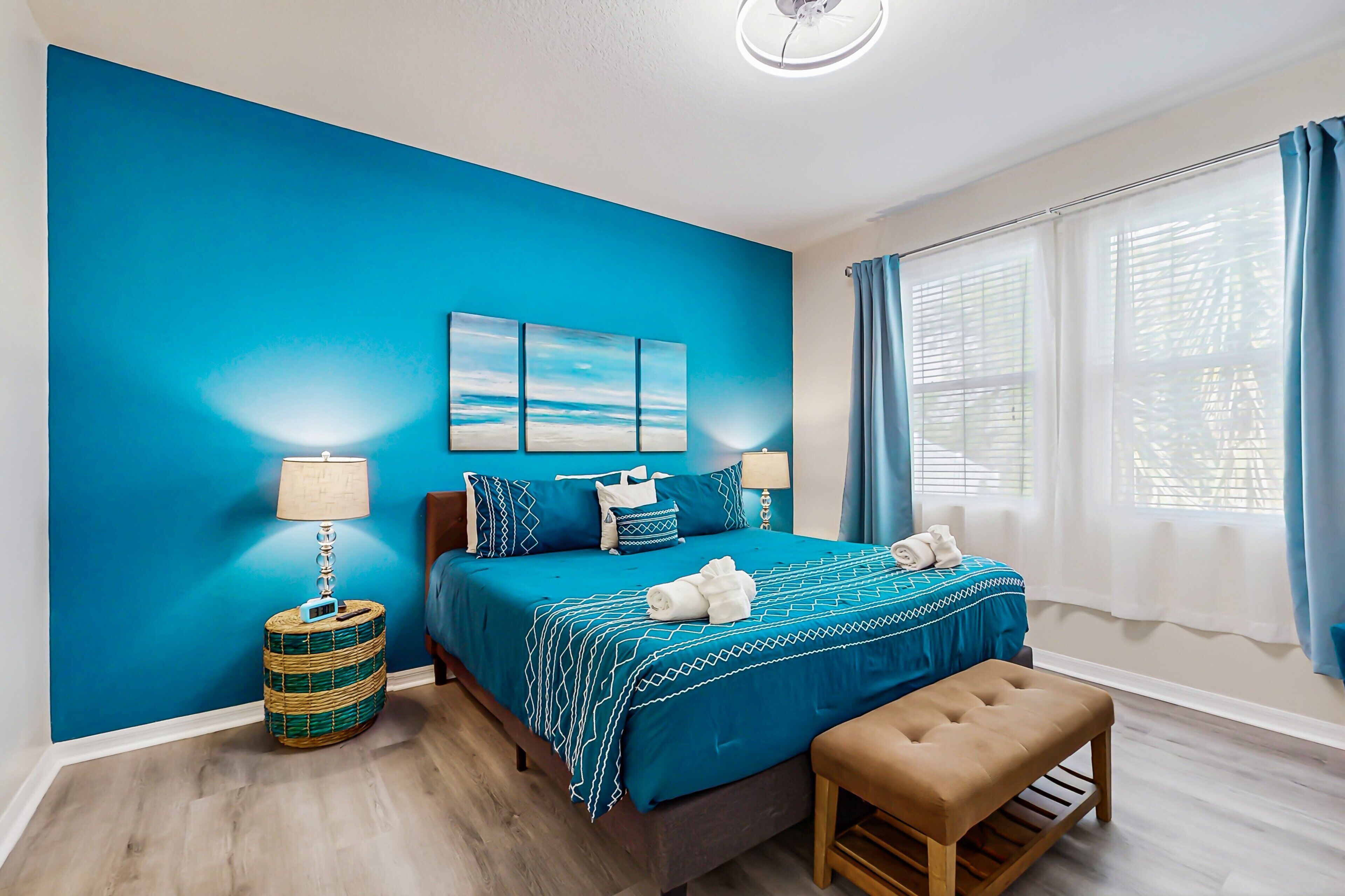 A bedroom with a blue wall, a bed with a blue bedspread, a bedside table with a lamp, a bench, and a window with curtains.