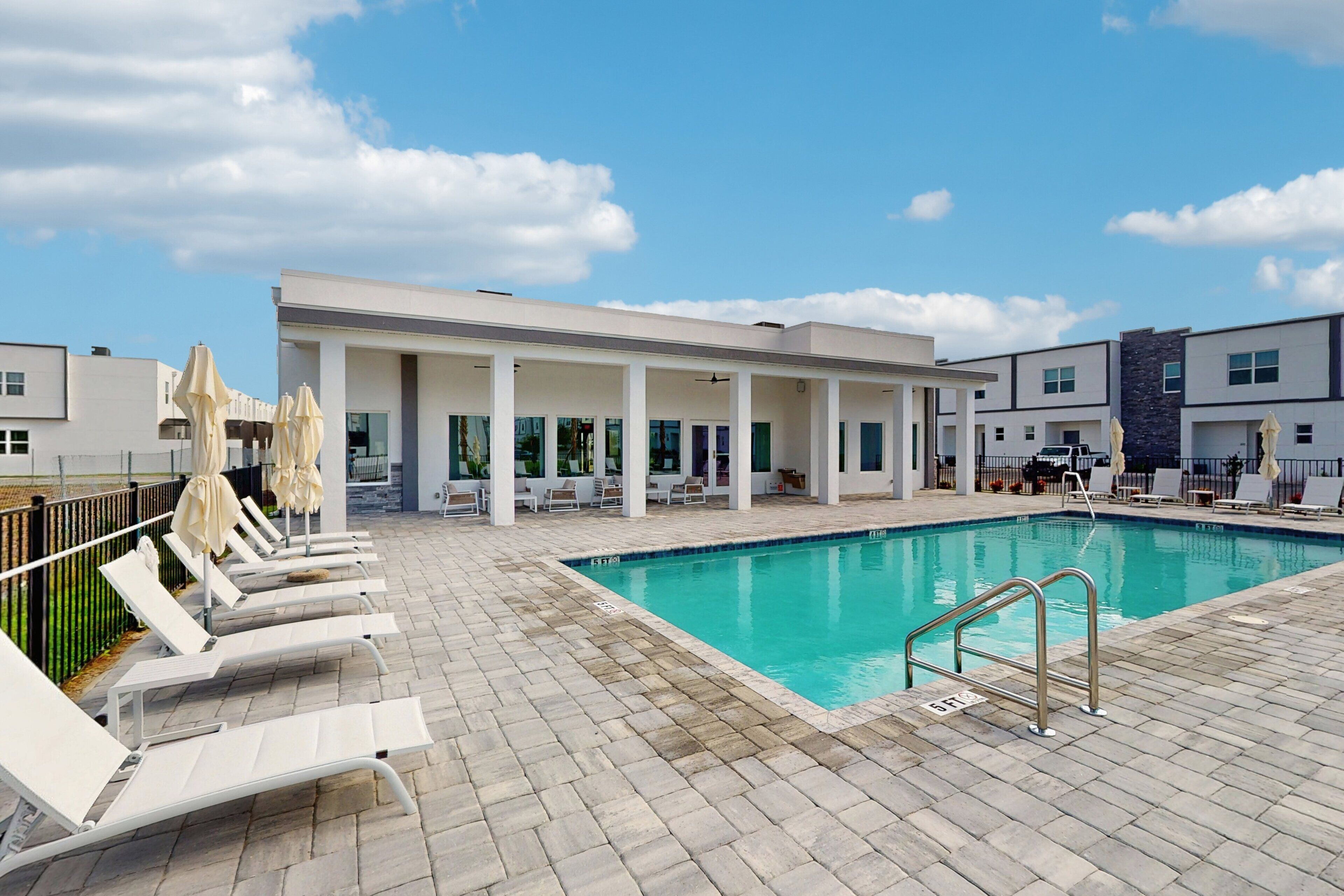 A swimming pool with lounge chairs and umbrellas, a covered seating area, and a modern building in the background.