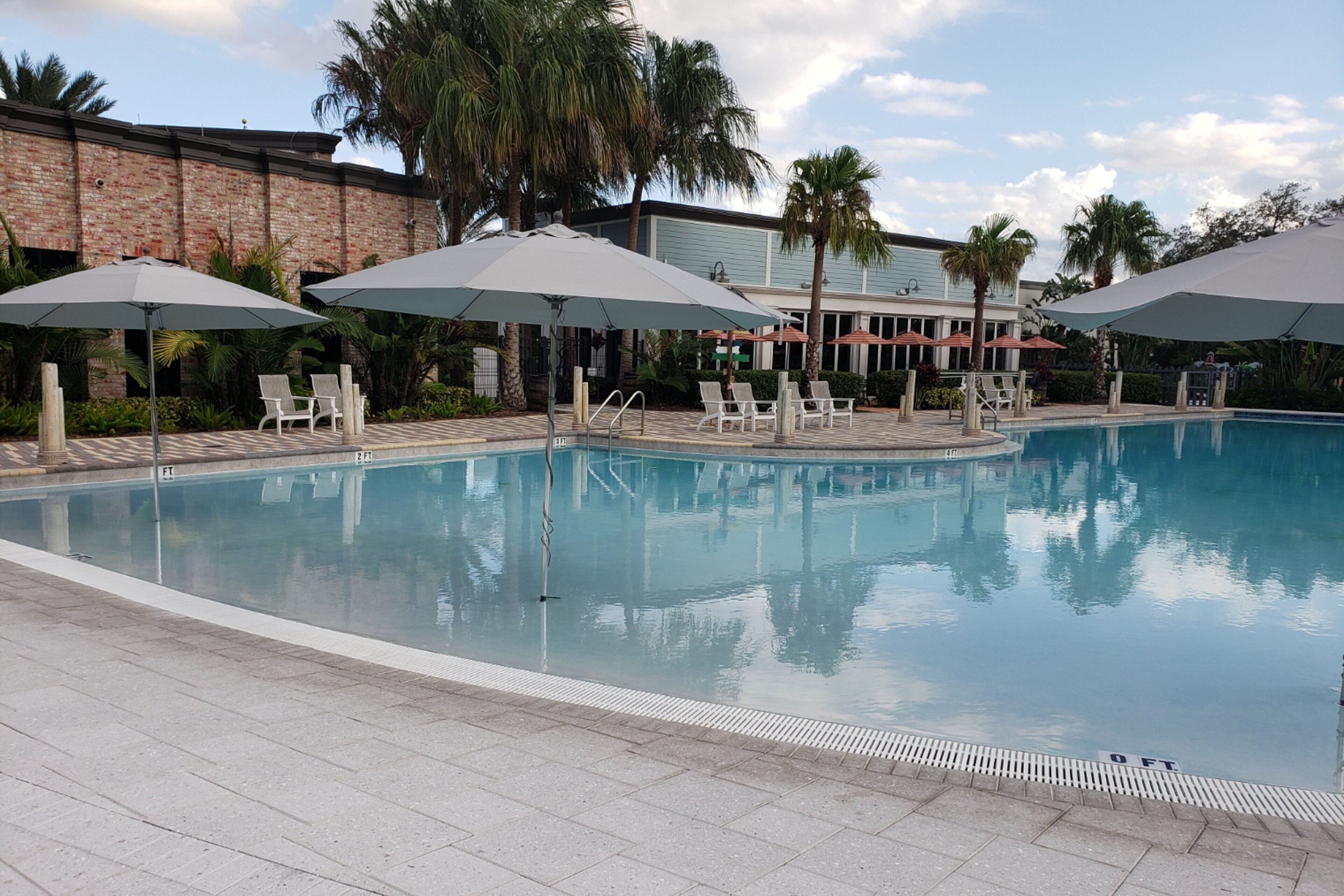 A swimming pool with lounge chairs and umbrellas, surrounded by palm trees and a brick building.