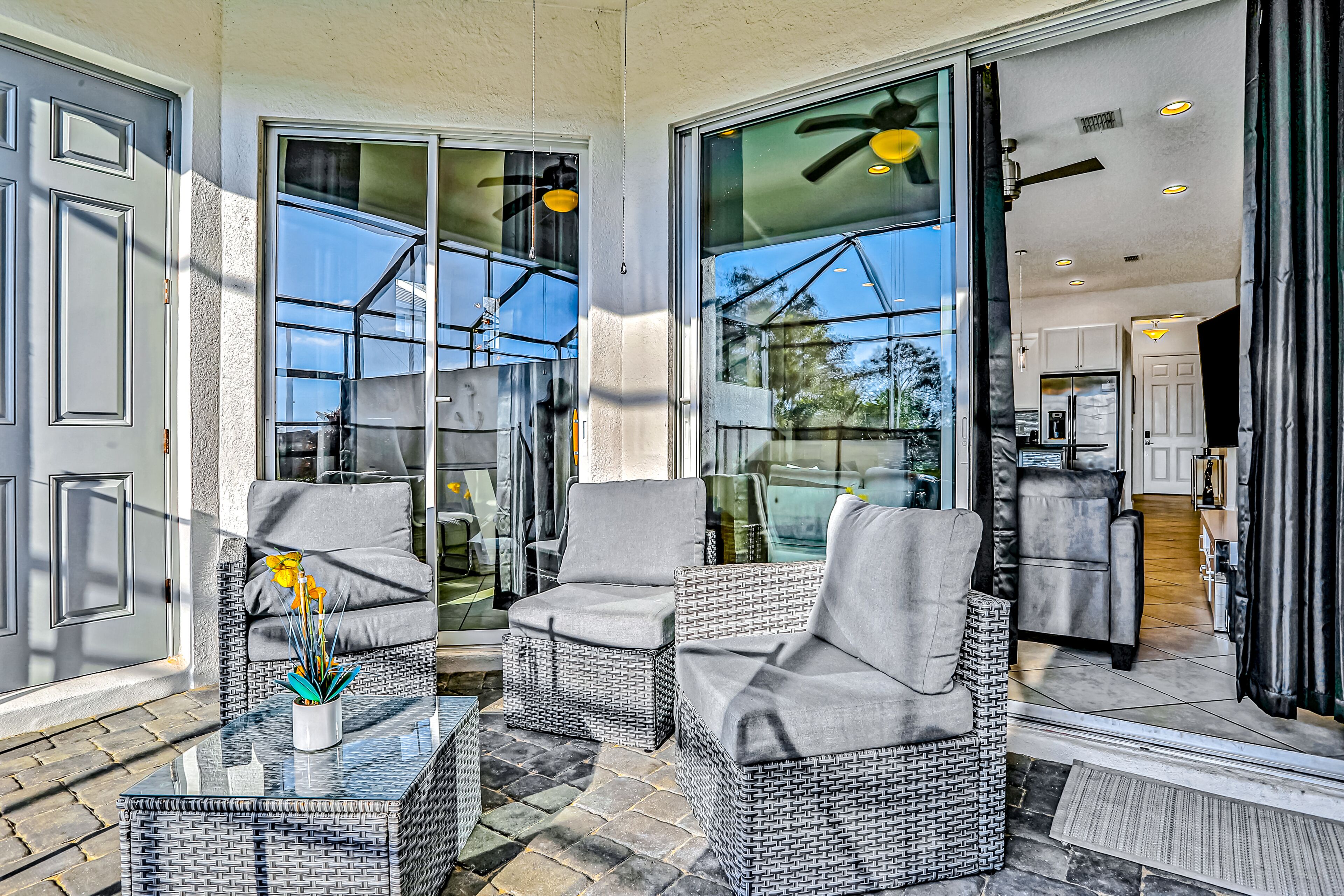 A sunlit patio with wicker furniture, a glass door leading to a kitchen, and a potted plant on a glass table.
