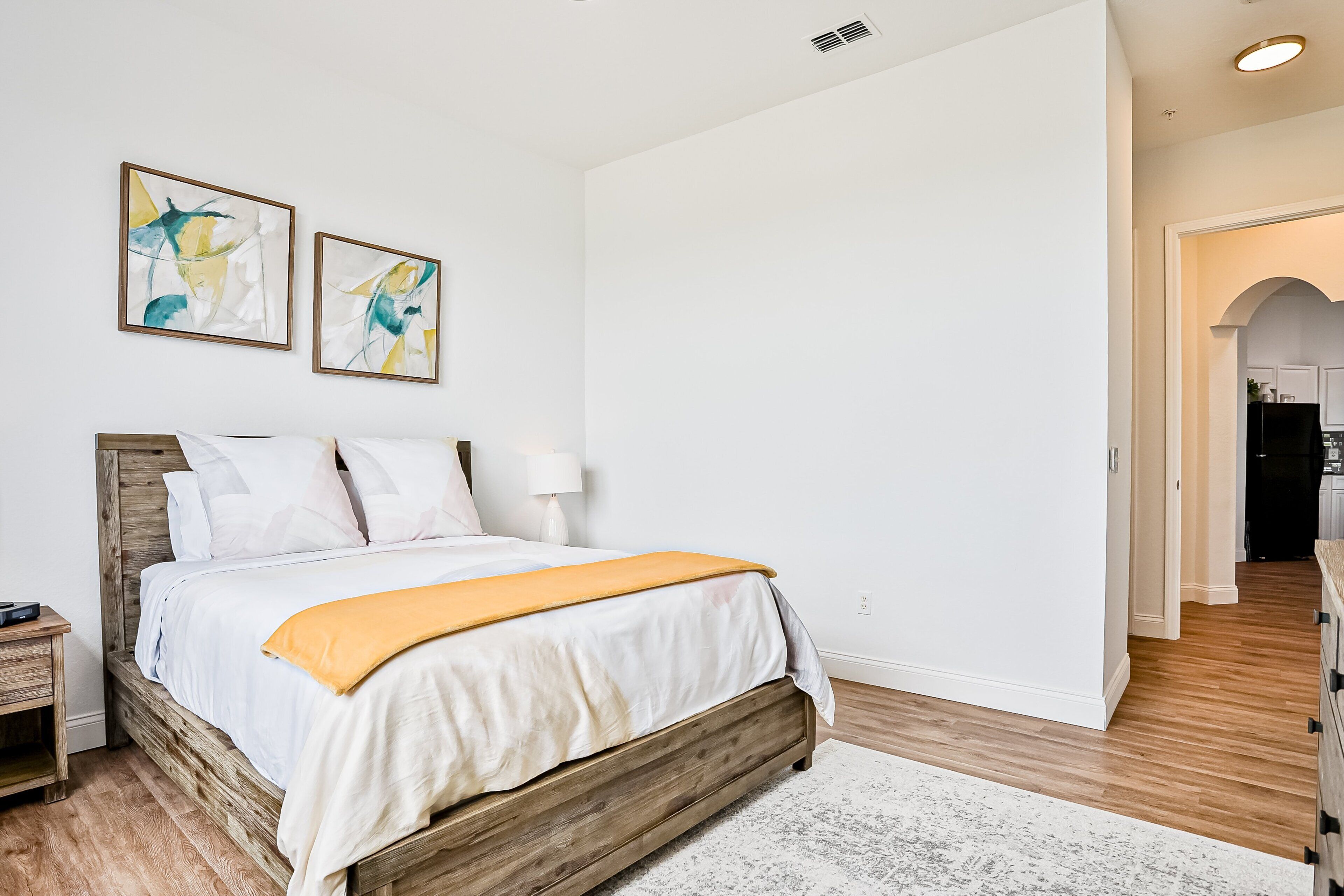 Camera da letto con un letto di legno, biancheria bianca, due dipinti astratti, un comodino e un corridoio che conduce a una cucina.