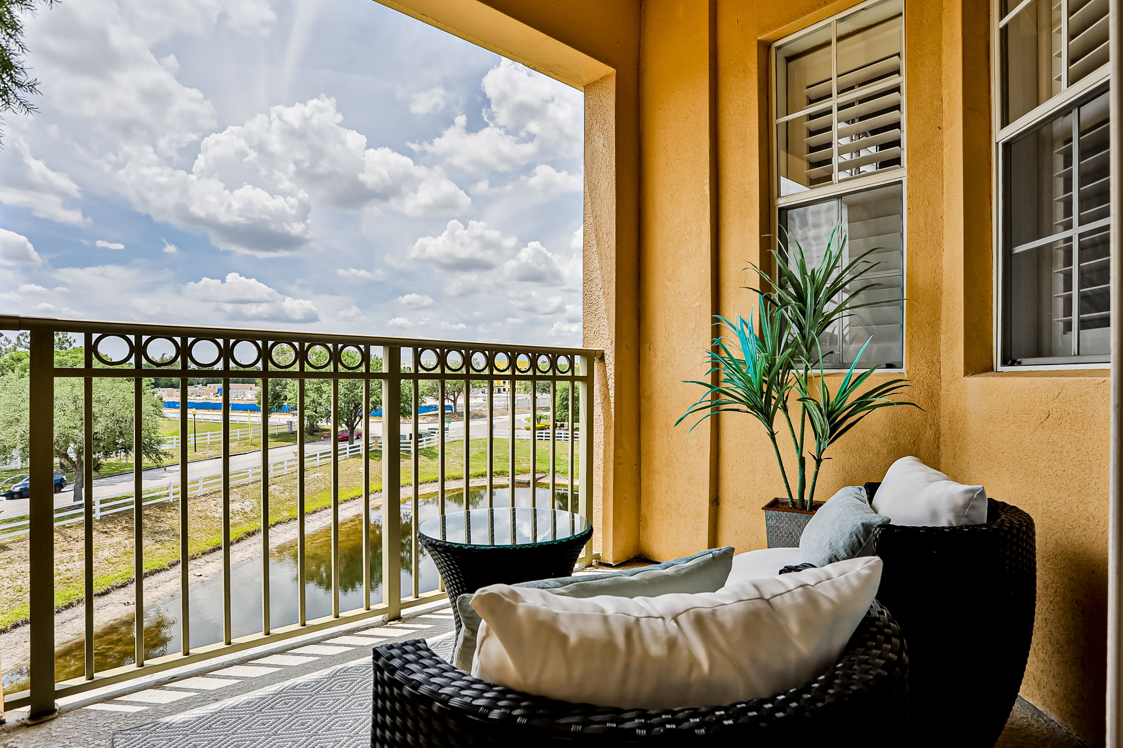 A balcony with a view of a road and a body of water, a wicker chair with cushions, and a potted plant.