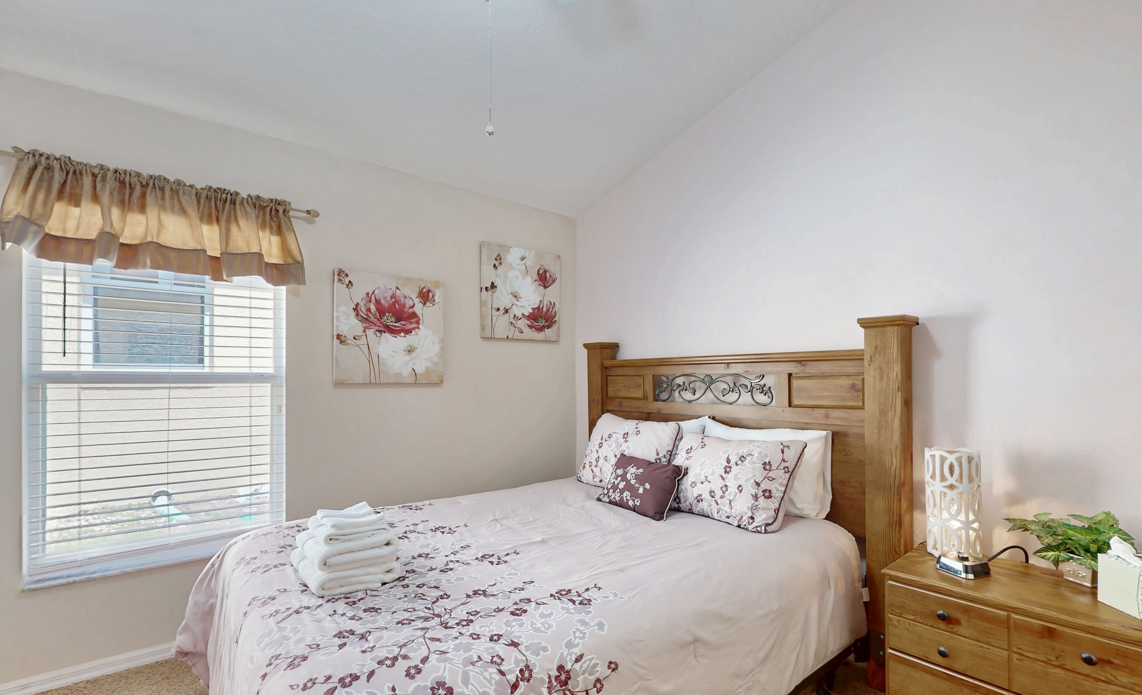 Una cama bien tendida con ropa de cama floral, un cabecero de madera y una mesita de noche con una lámpara y una planta.