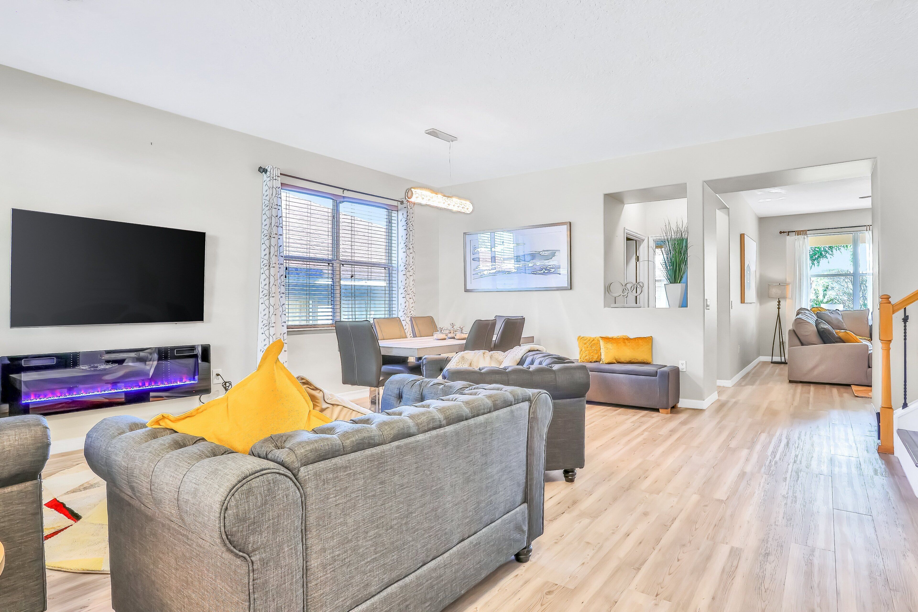 A modern living room with a flat-screen TV, grey sofas, a dining area, and a staircase leading to another level.