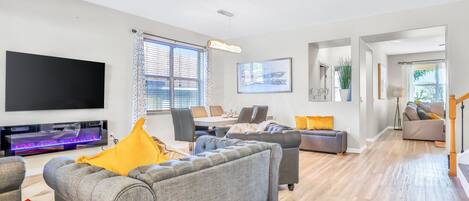A modern living room with a flat-screen TV, grey sofas, a dining area, and a staircase leading to another level.