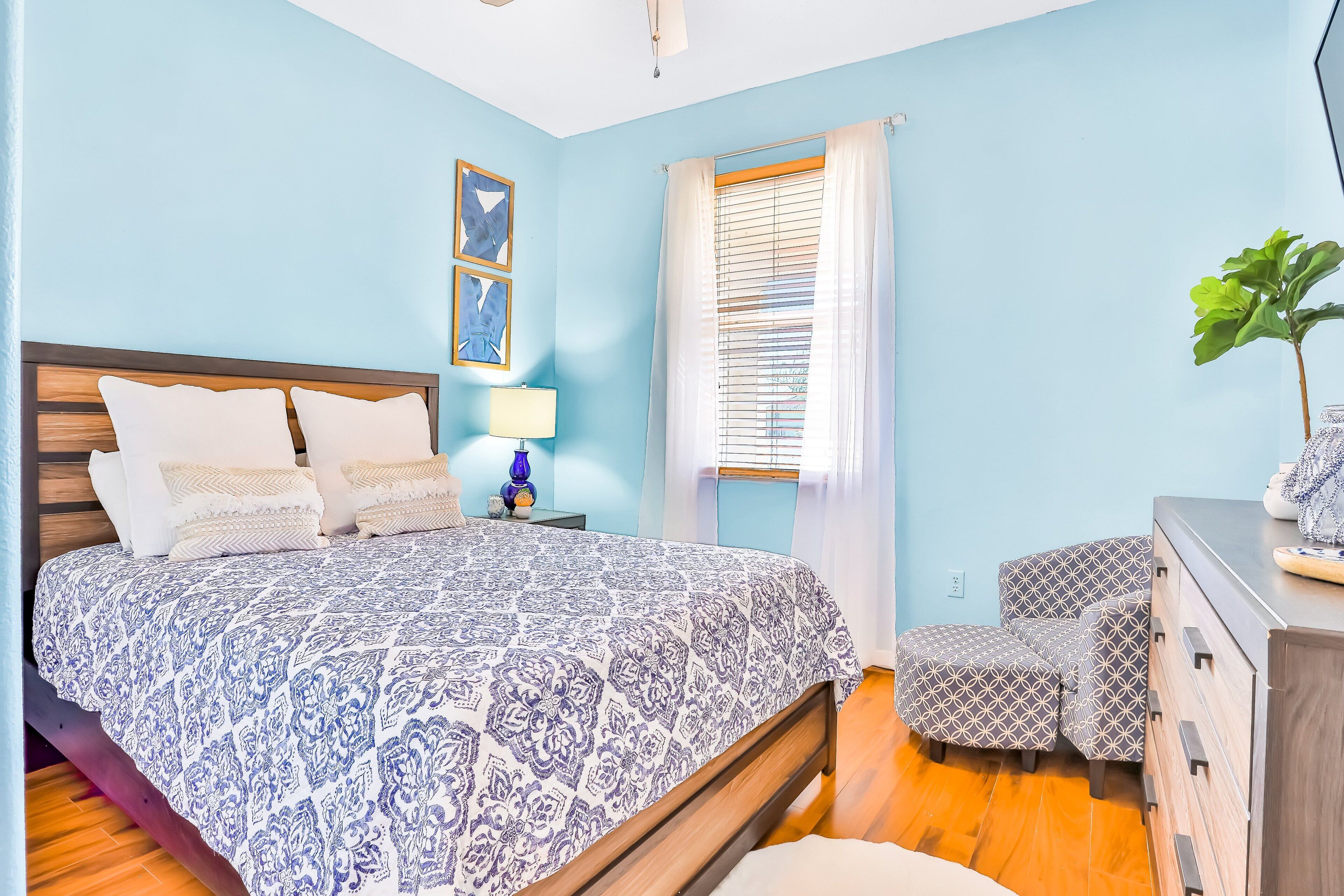 A bedroom with a bed, bedside table, lamp, window with blinds, and a dresser.