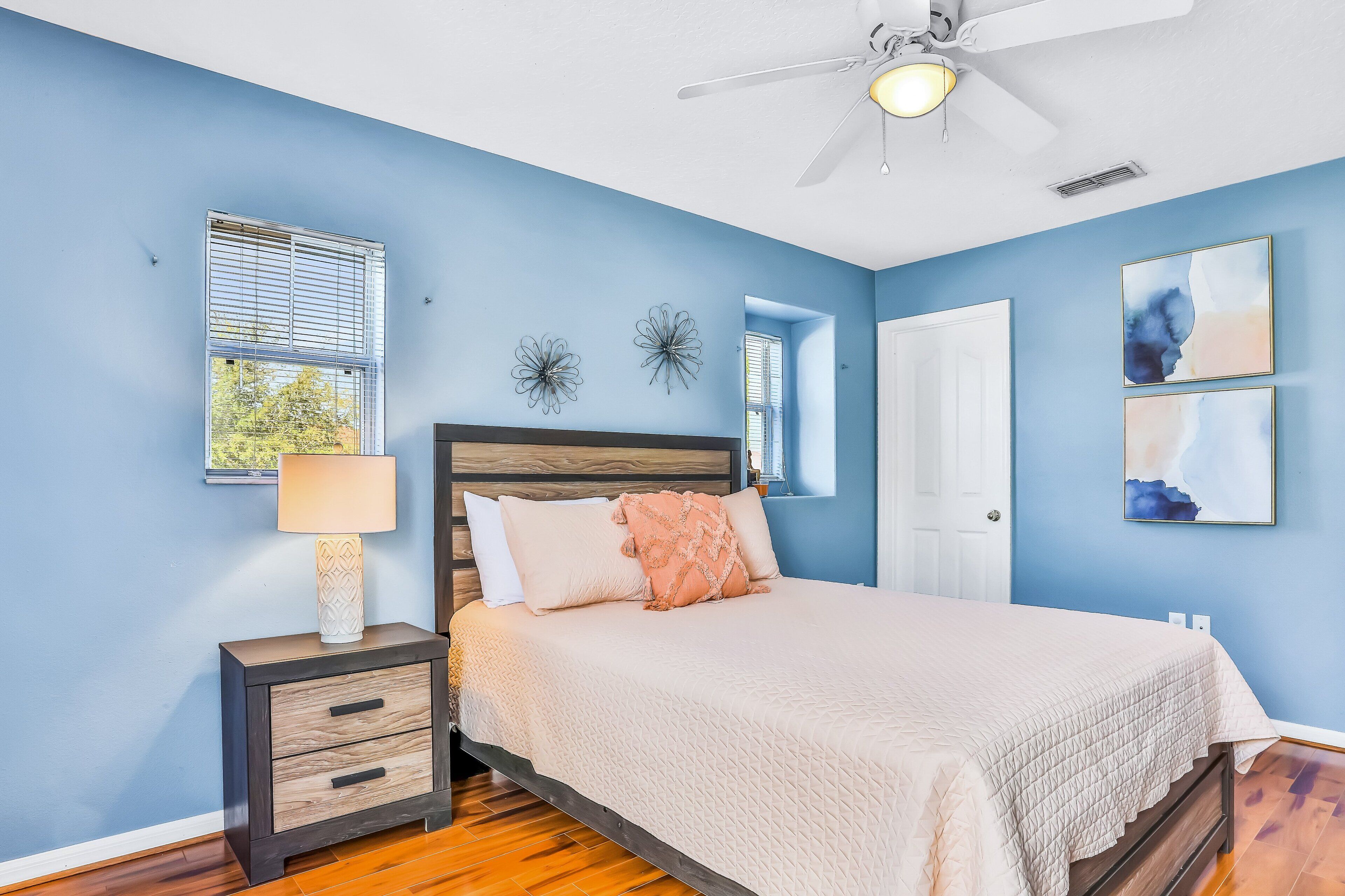 Un dormitorio con cama, mesita de noche, decoración en la pared, ventilador de techo y ventana con persianas.