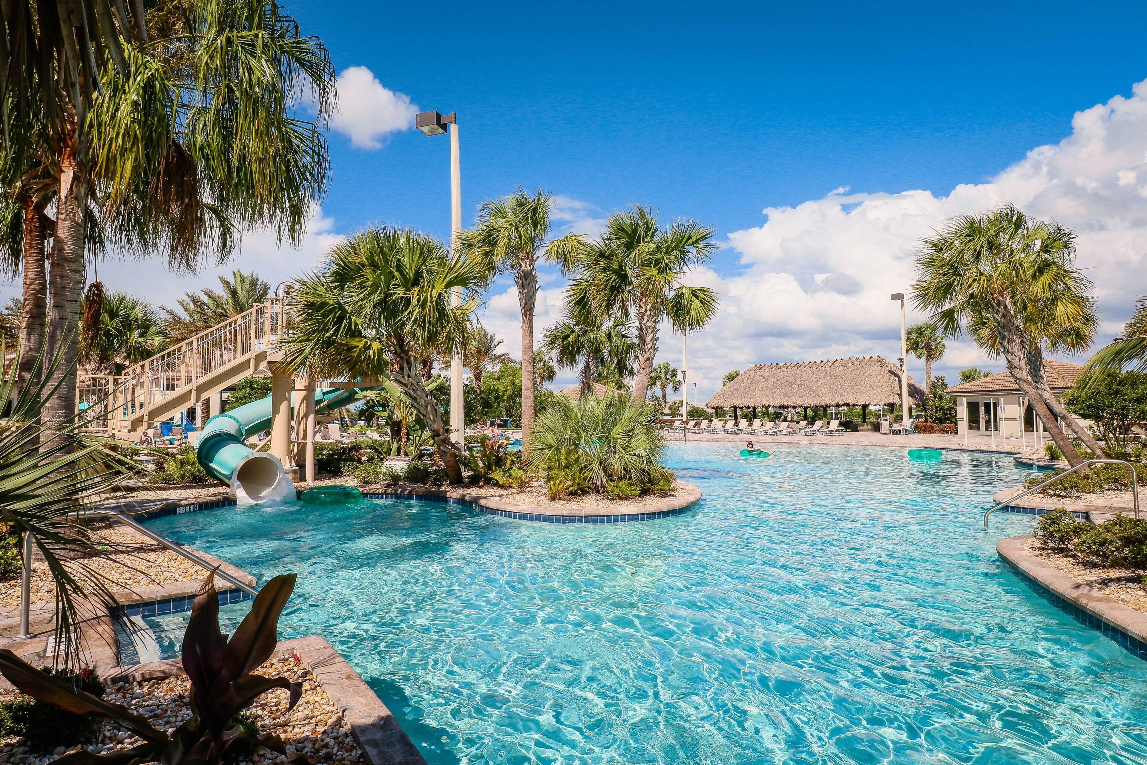 A swimming pool with a slide, palm trees, and a building with a thatched roof.
