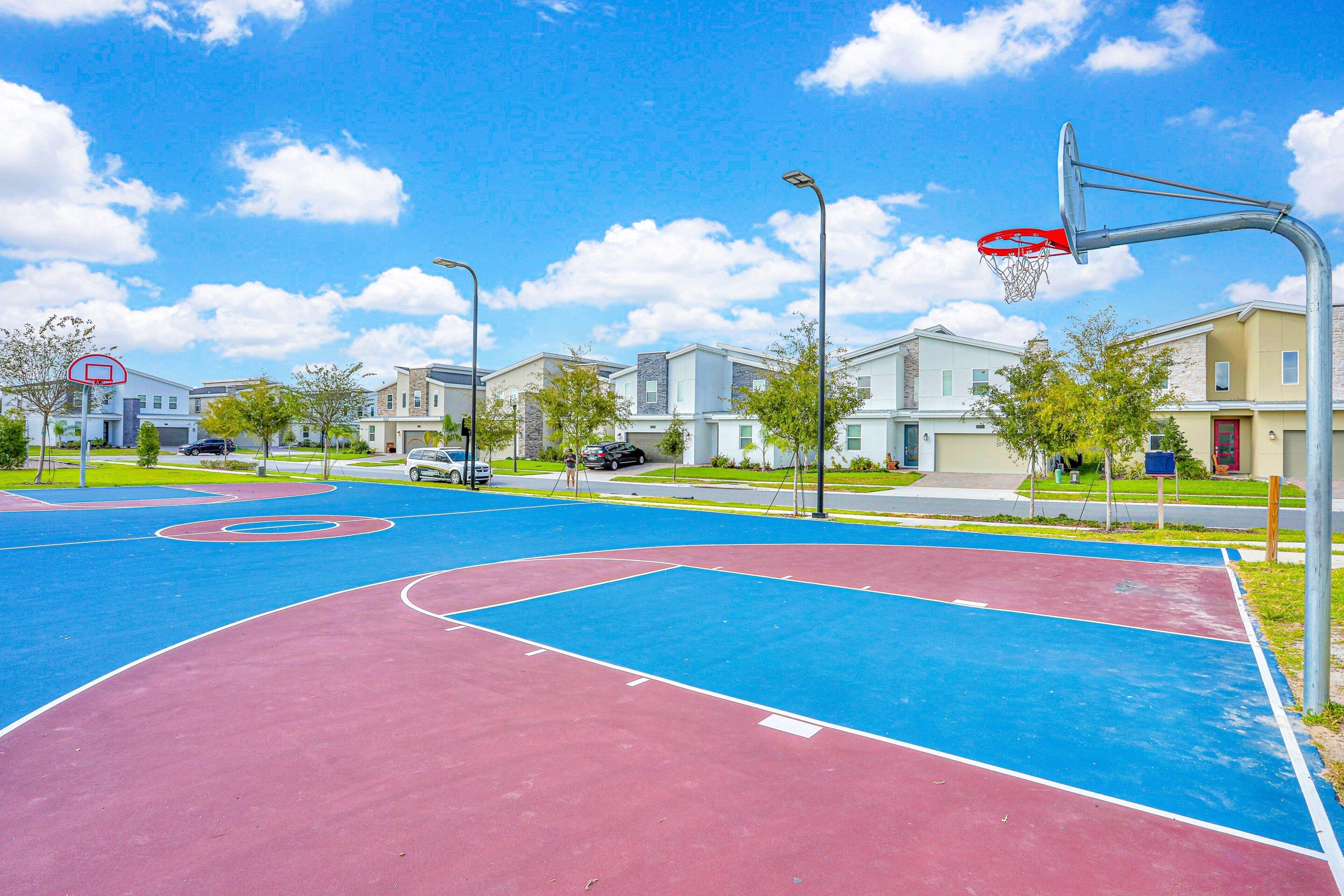 Un terrain de basket en plein air, avec une surface de jeu rouge et bleue, entouré d’immeubles d’habitation.